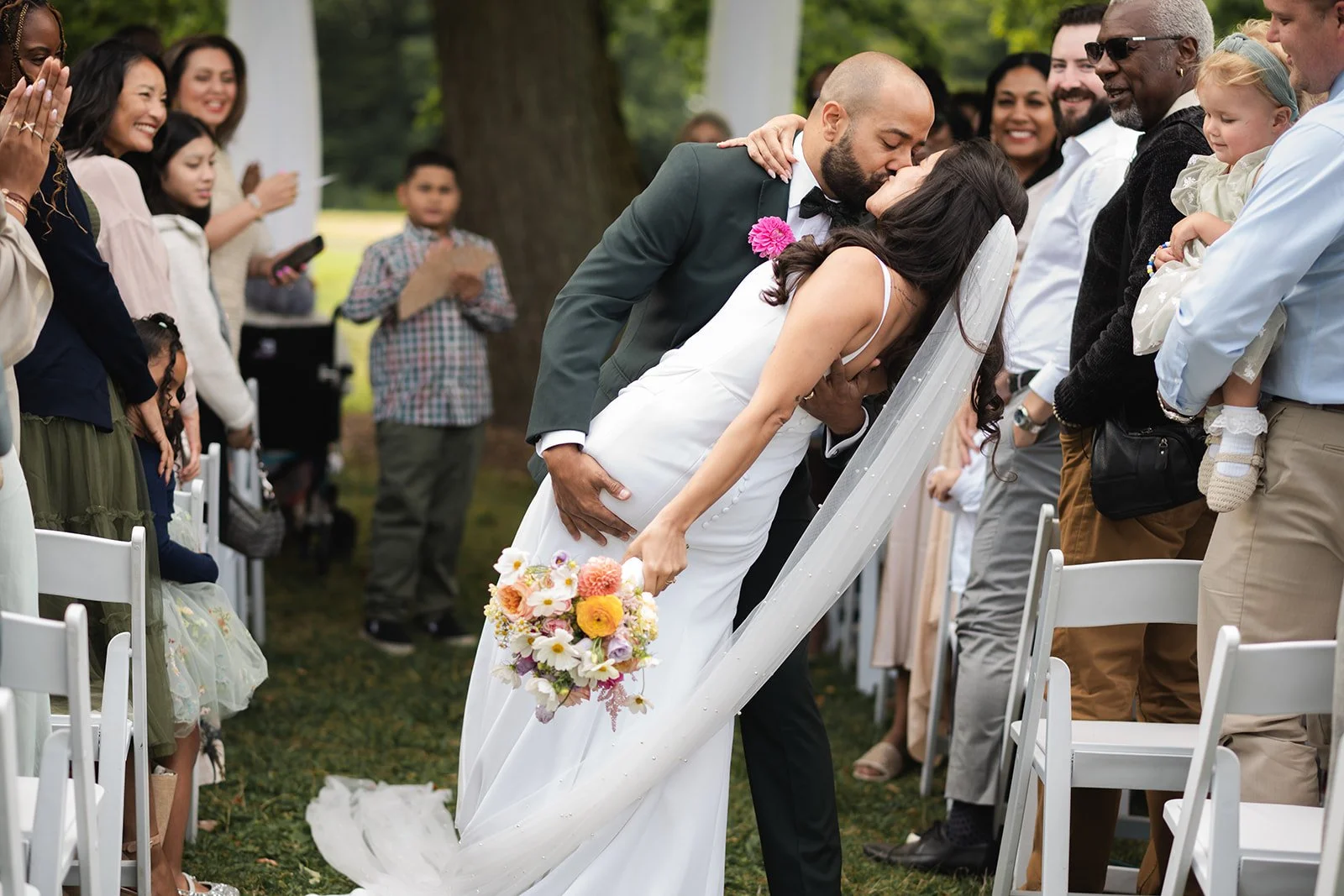 A wedding couple shares a kiss during their outdoor ceremony surrounded by guests, with the bride holding a colorful bouquet of flowers.