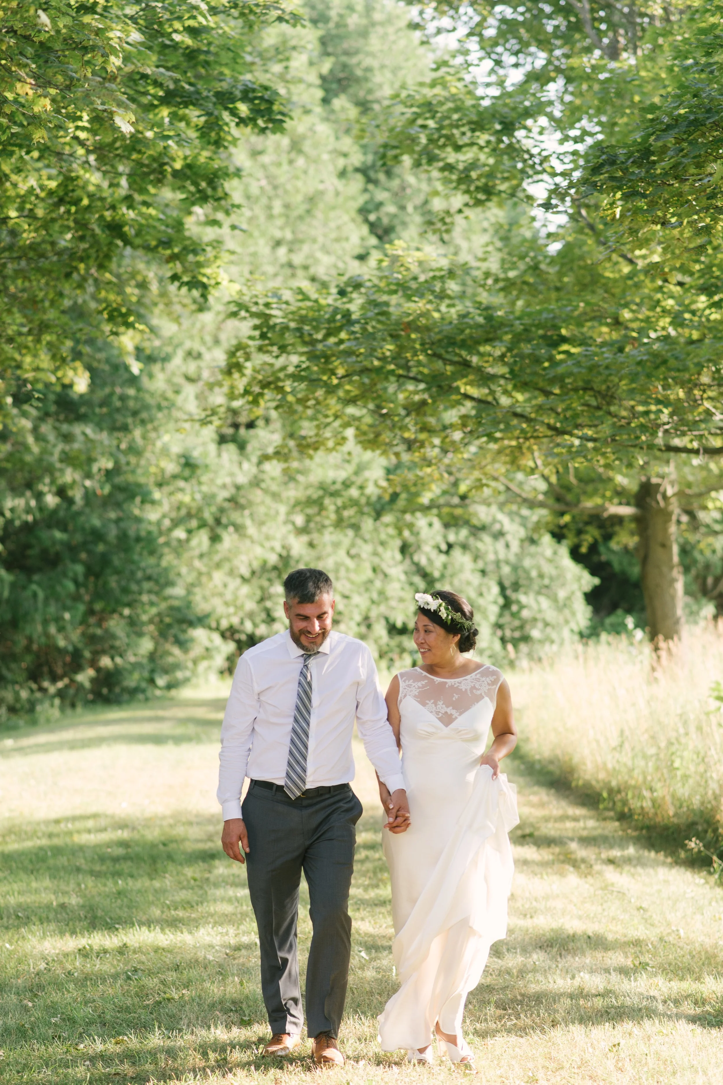 A bride and groom walking hand in hand in a park with lush green trees, one woman in a wedding dress with a floral headband and one man in a white shirt and tie.