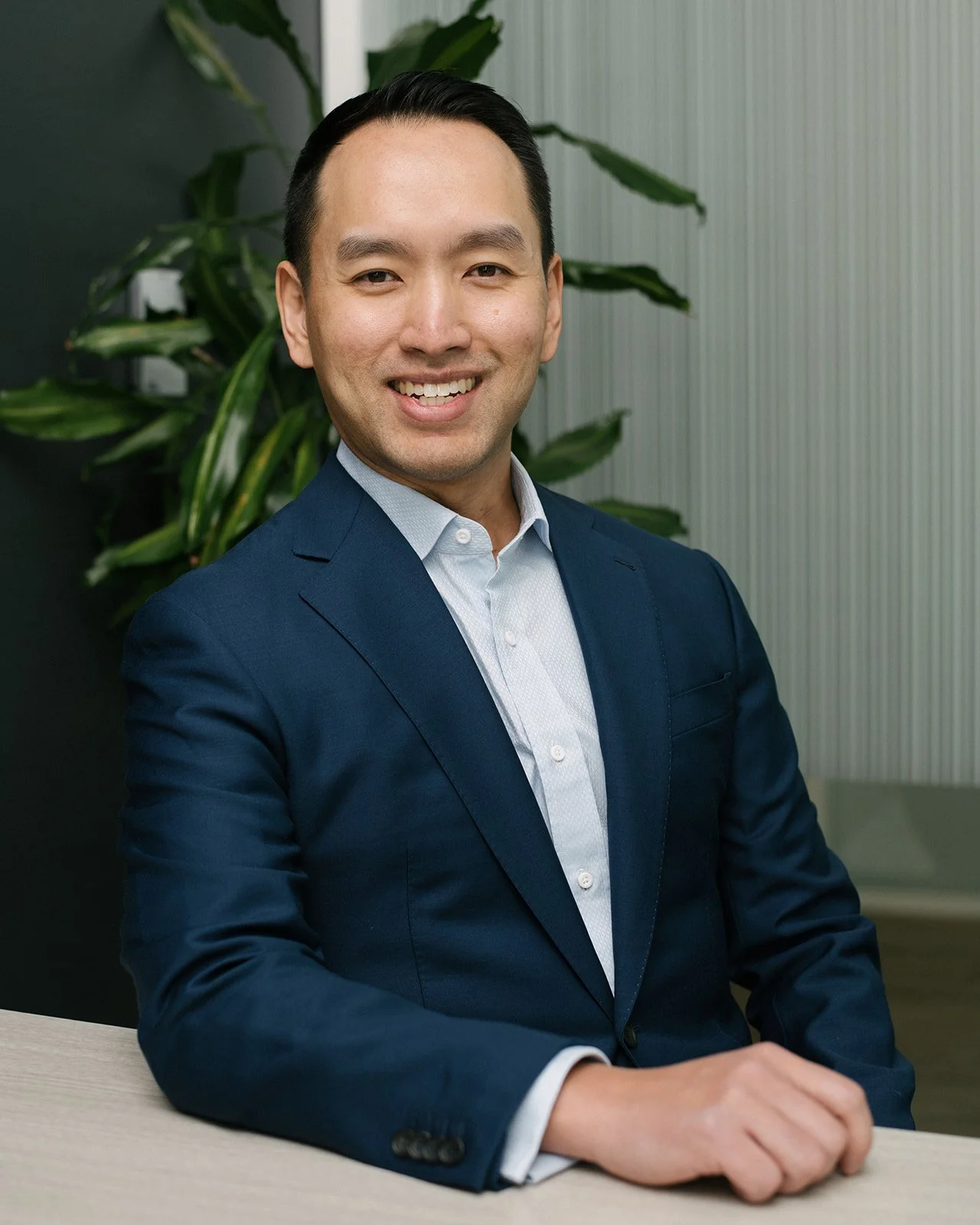 A corporate headshot of a man with dark hair smiling, wearing a blue suit and light blue shirt, sitting at a desk in an office setting with green plants in the background.