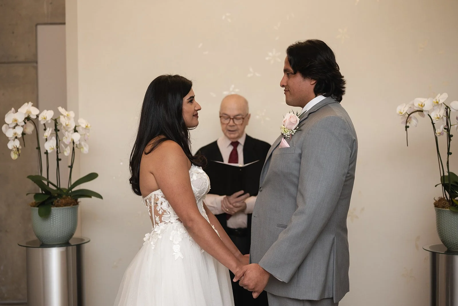 A bride and groom holding hands during their wedding ceremony, facing each other, with an officiant reading from a book behind them. There are white orchids on either side of the couple.