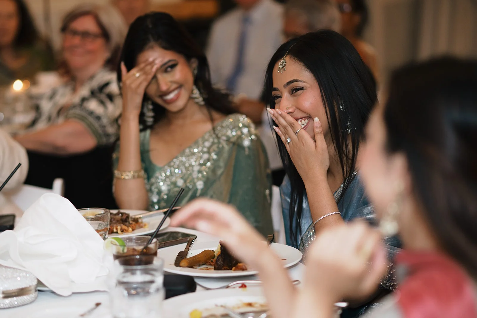 Two women wearing traditional Indian attire sitting at a table, sharing a joyful moment and laughing during a wedding celebration at The Broadview Hotel in Toronto.