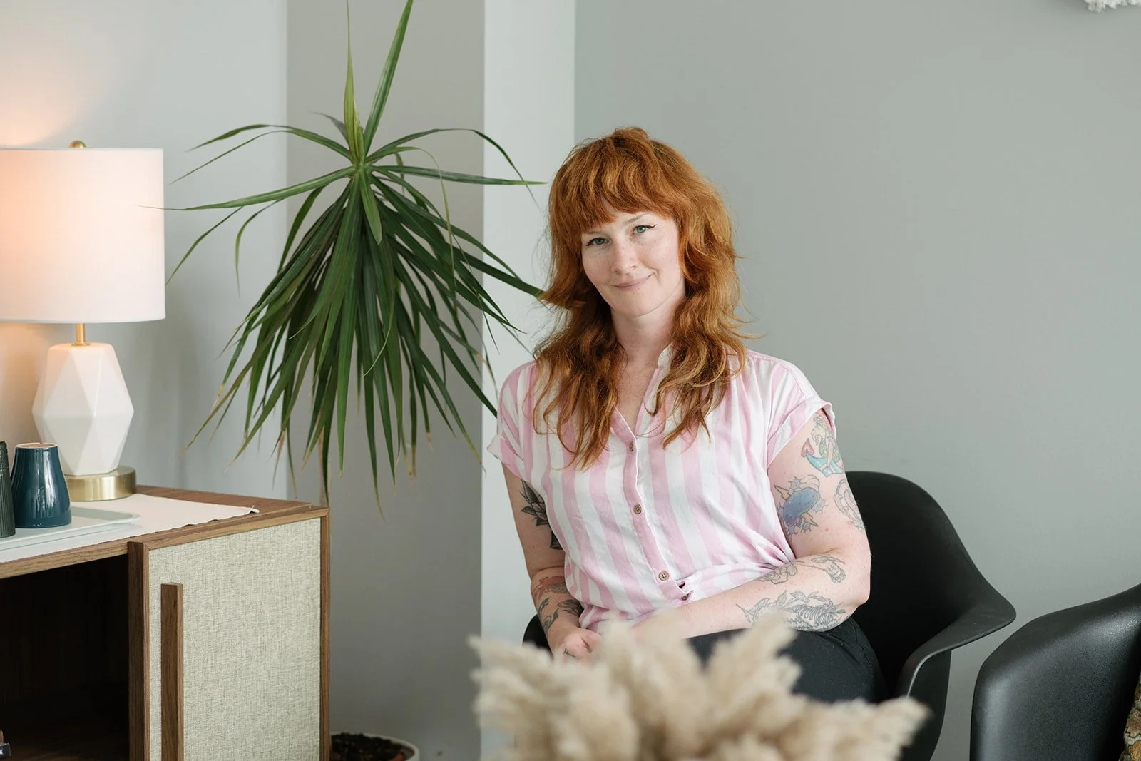 A person with red hair, wearing a pink and white striped shirt, sitting in a modern looking waiting area at Origin Wellness in Toronto. 