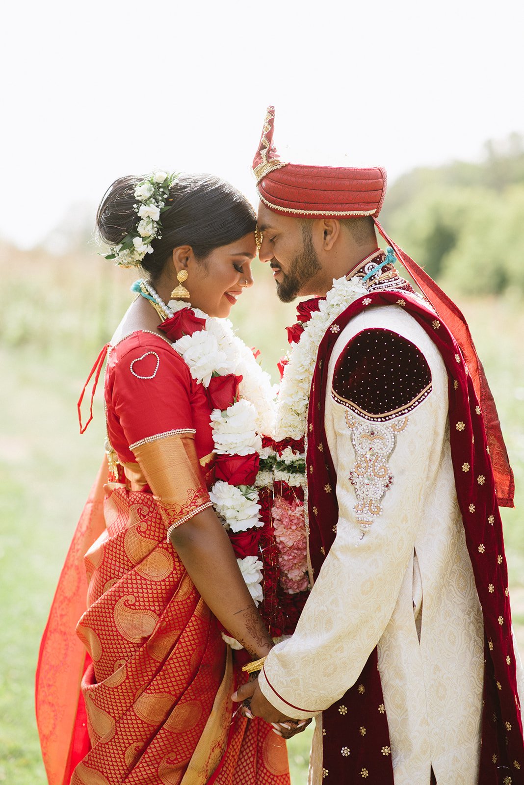 An Indian couple in wedding Attire holding hands and touching foreheads with eyes closed, outdoors during daytime.