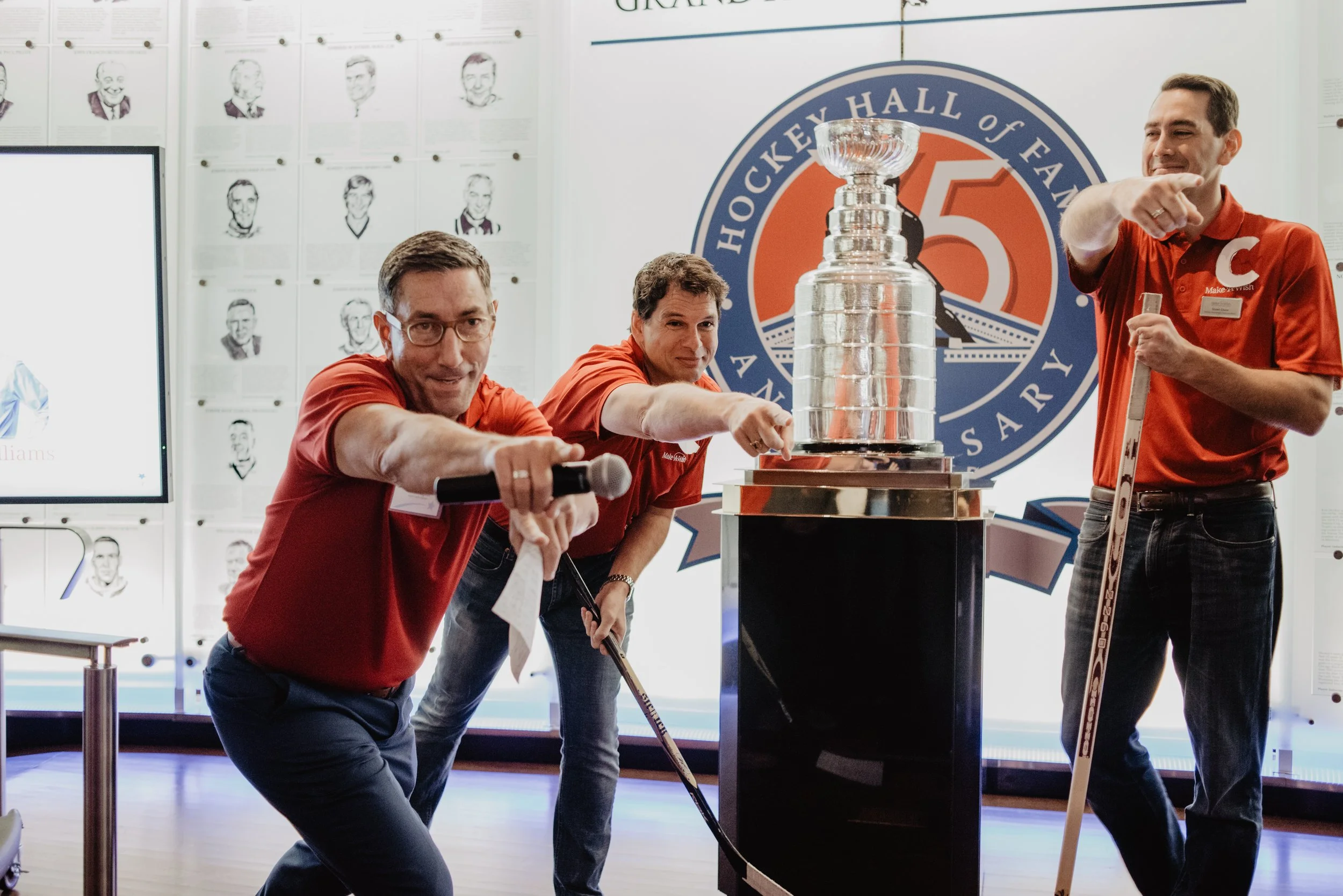 Three men in red shirts with a hockey logo are posing with the Stanley Cup in an Exhibition Hall. The man on the left is holding a microphone, the man in the middle points at the trophy, and the man on the right holds a hockey stick. Behind them is a