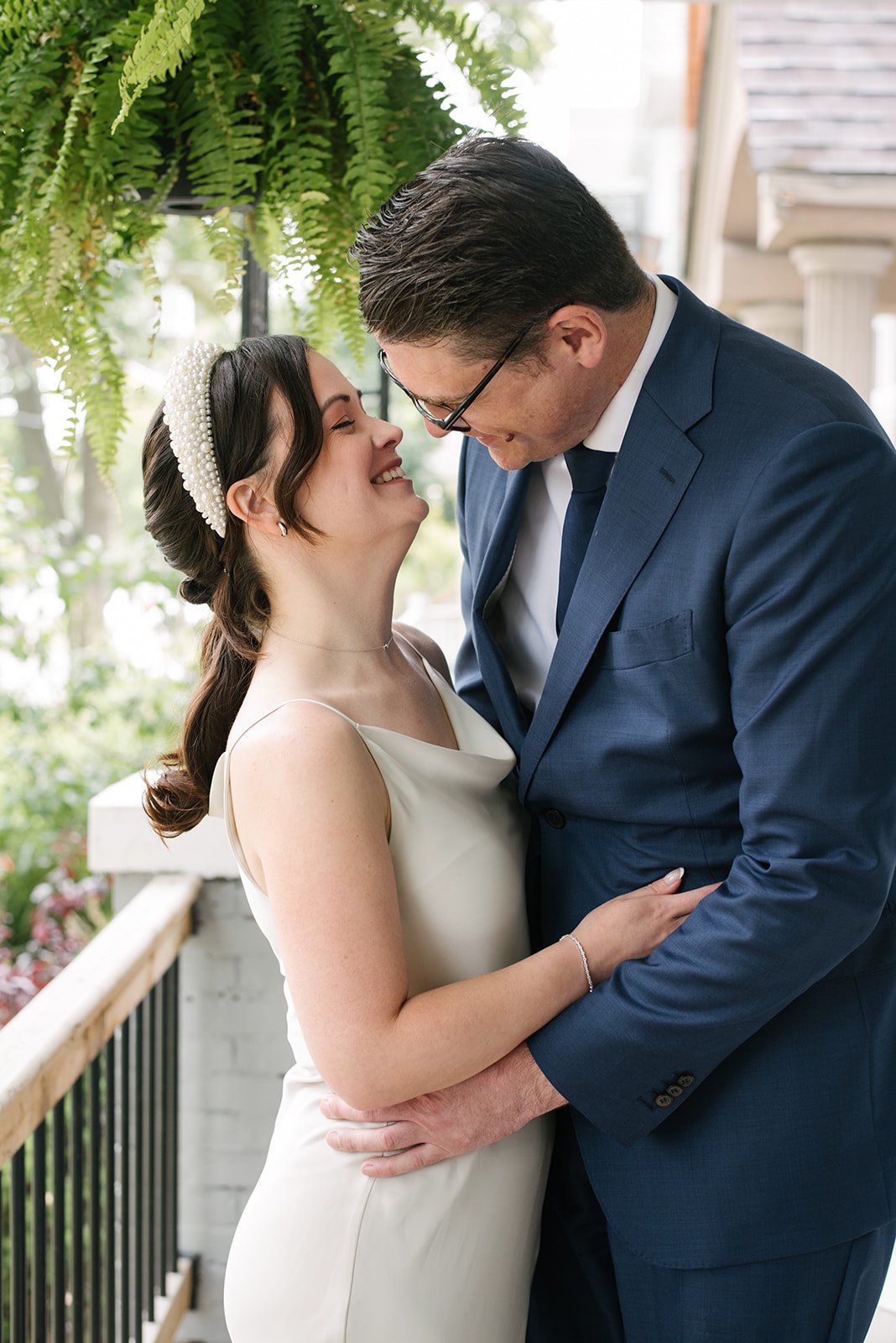 A bride and groom sharing a joyful moment indoors with green plants in the background. The bride is wearing a white dress and a pearl headband, and the groom is in a blue suit and glasses, with their foreheads touching and smiling.