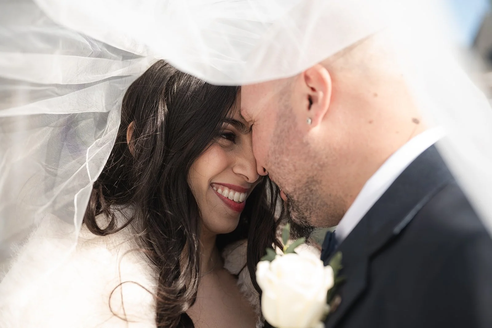 A bride and groom face each other under a veil, smiling and touching foreheads, after getting married at Toronto City Hall. 