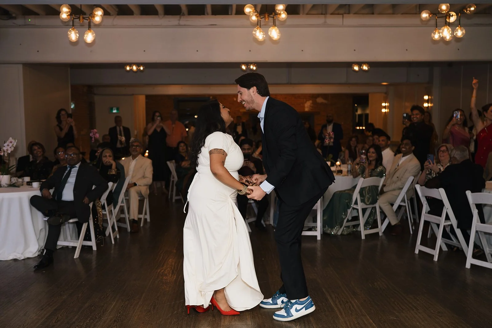 A couple sharing their first dance at a wedding reception in the Broadview Hotel in Toronto, surrounded by seated guests and dim lighting, with the man wearing a suit and sneakers and the woman in a white dress and red heels.