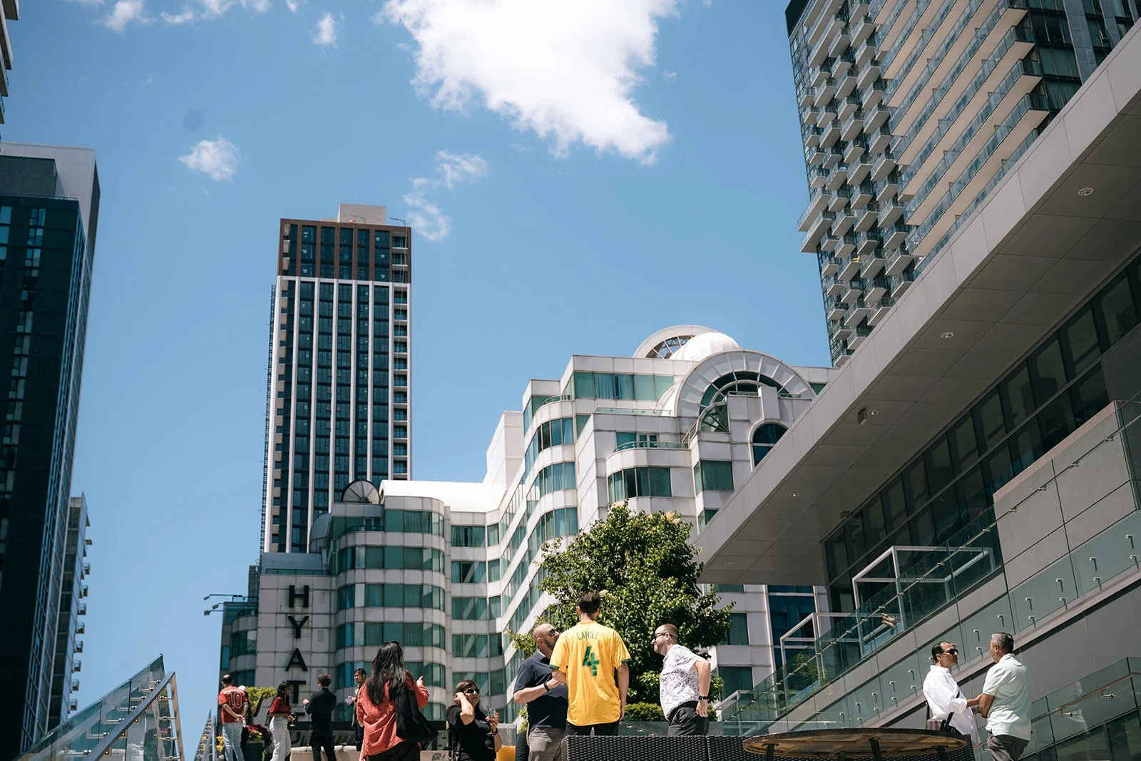 People gathered outdoors at a corporate event on a city terrace with tall modern buildings in the background under a blue sky.