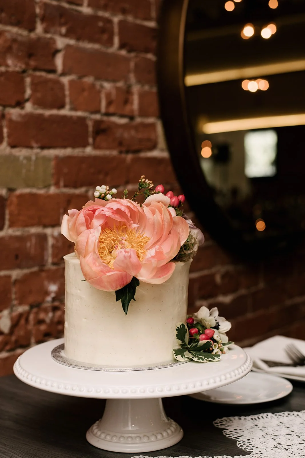 A white, wedding cake with a large pink flower and small berries on top, placed on a white cake stand with a decorative edge, set against a brick wall with a round mirror and bokeh light reflections.