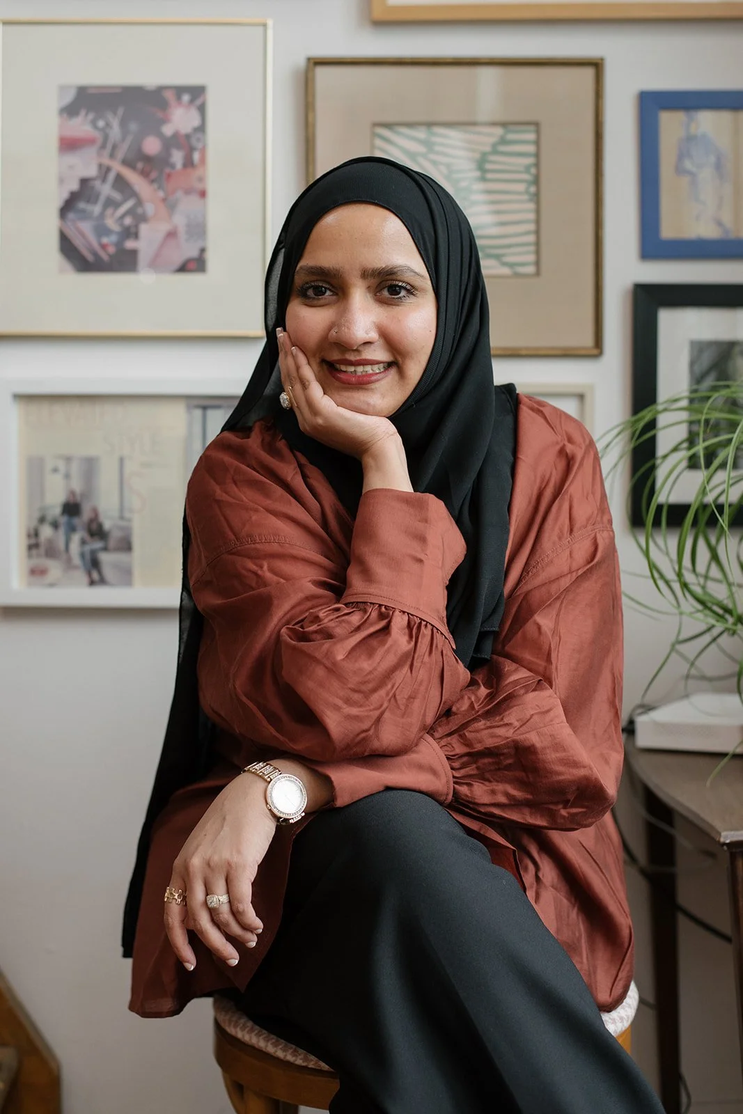 A woman wearing a black hijab and a rust-colored shirt, sitting on a chair with her chin resting on her hand, in front of a wall with framed artwork and a plant nearby.