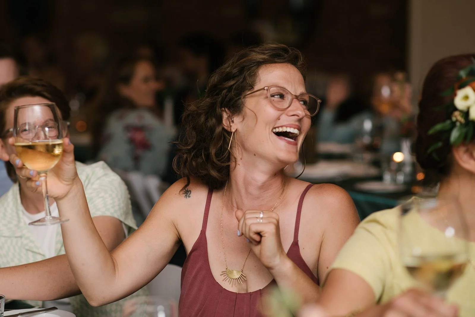 A woman with curly hair, glasses, and earrings, wearing a red dress, laughing and holding a glass of white wine at a wedding reception at The Broadview Hotel.
