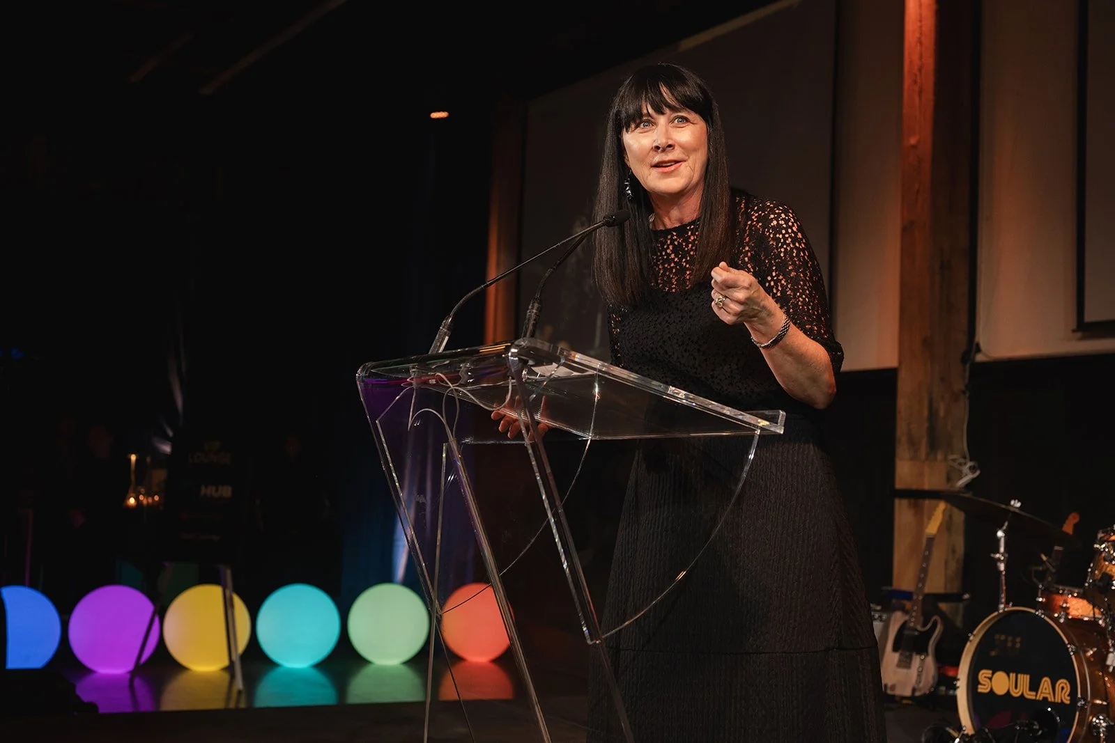 A woman with dark hair and bangs speaking at a clear podium with a microphone, wearing a black lace top. She is gesturing with her right hand. The background includes colorful illuminated spheres and musical instruments like drums and guitars.
