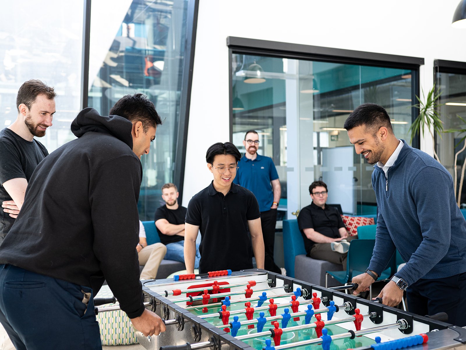 Group of men smiling and playing foosball in a modern office lounge area with large windows, some sitting on sofas, others standing around the foosball table.