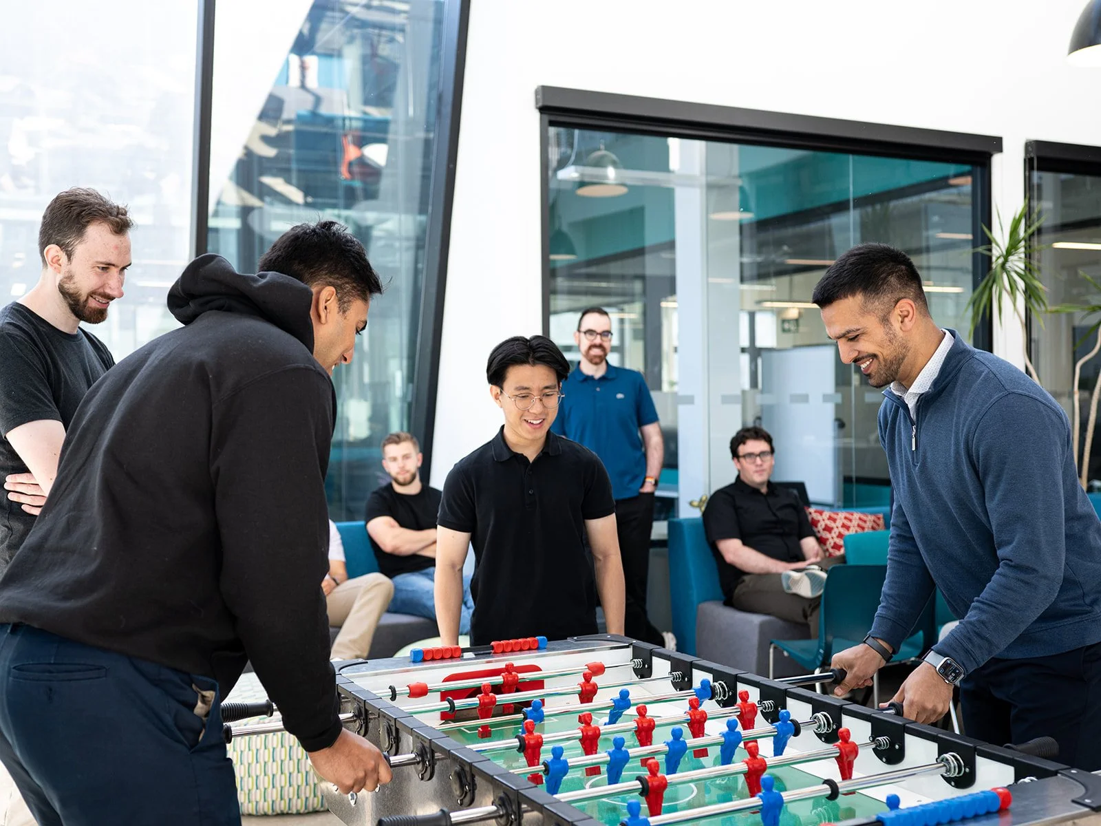 Group of men smiling and playing foosball in a modern office lounge area with large windows, some sitting on sofas, others standing around the foosball table.