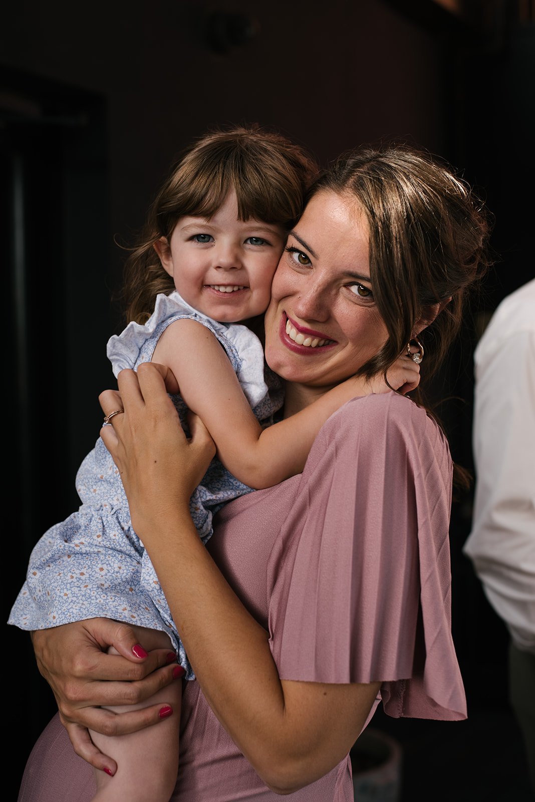 A woman with brown hair in a pink dress holding a young girl with brown hair in a light blue dress, both smiling and hugging each other, against a dark background.