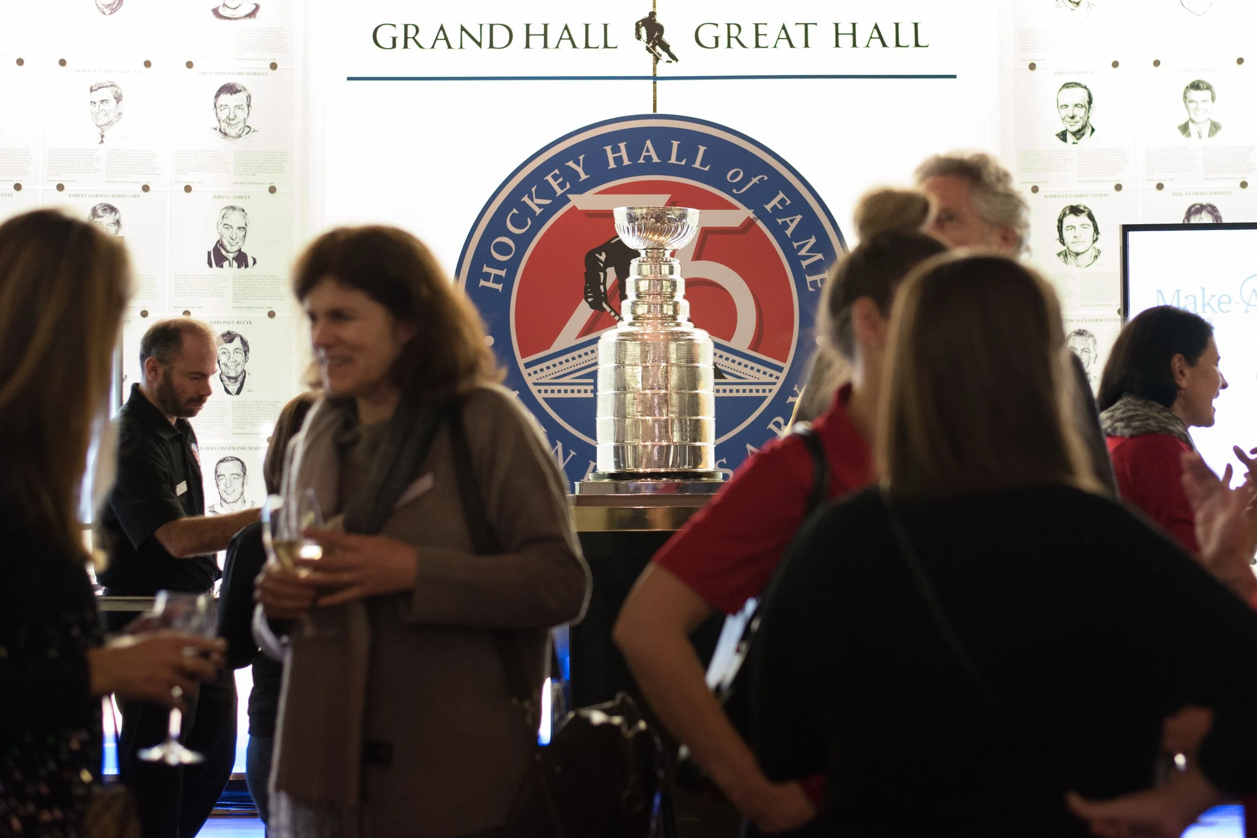 Celebration event at the Hockey Hall of Fame with a large replica of the Stanley Cup trophy, surrounded by people socializing and enjoying drinks, under a backdrop featuring the Hockey Hall of Fame logo and a sign reading 'Grand Hall Great Hall'.