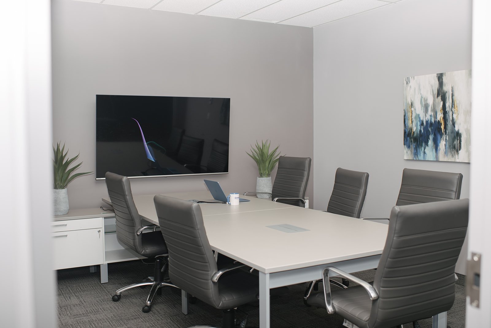 An empty modern conference room with a rectangular table, six black leather chairs, a large wall-mounted screen, two potted plants, a laptop, a coffee mug, and abstract artwork on the wall.