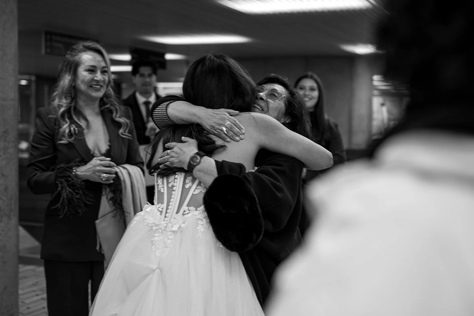 A woman in a wedding dress hugging an older woman, who appears emotional, surrounded by smiling people at Toronto City Hall