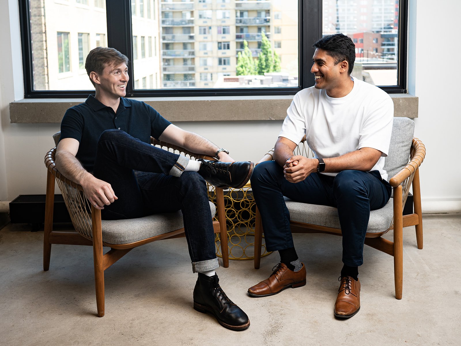 Two co-owners of Terminal sitting on chairs and smiling at each other in a bright room with large windows overlooking buildings.