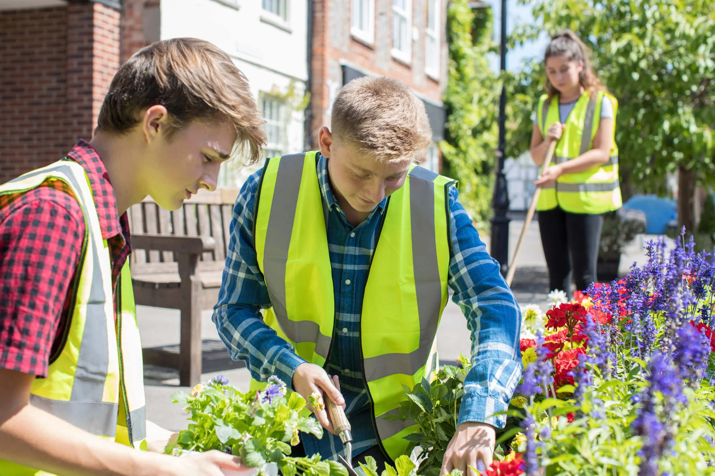 Three young volunteers in yellow safety vests planting flowers in an outdoor park or garden area on a sunny day.
