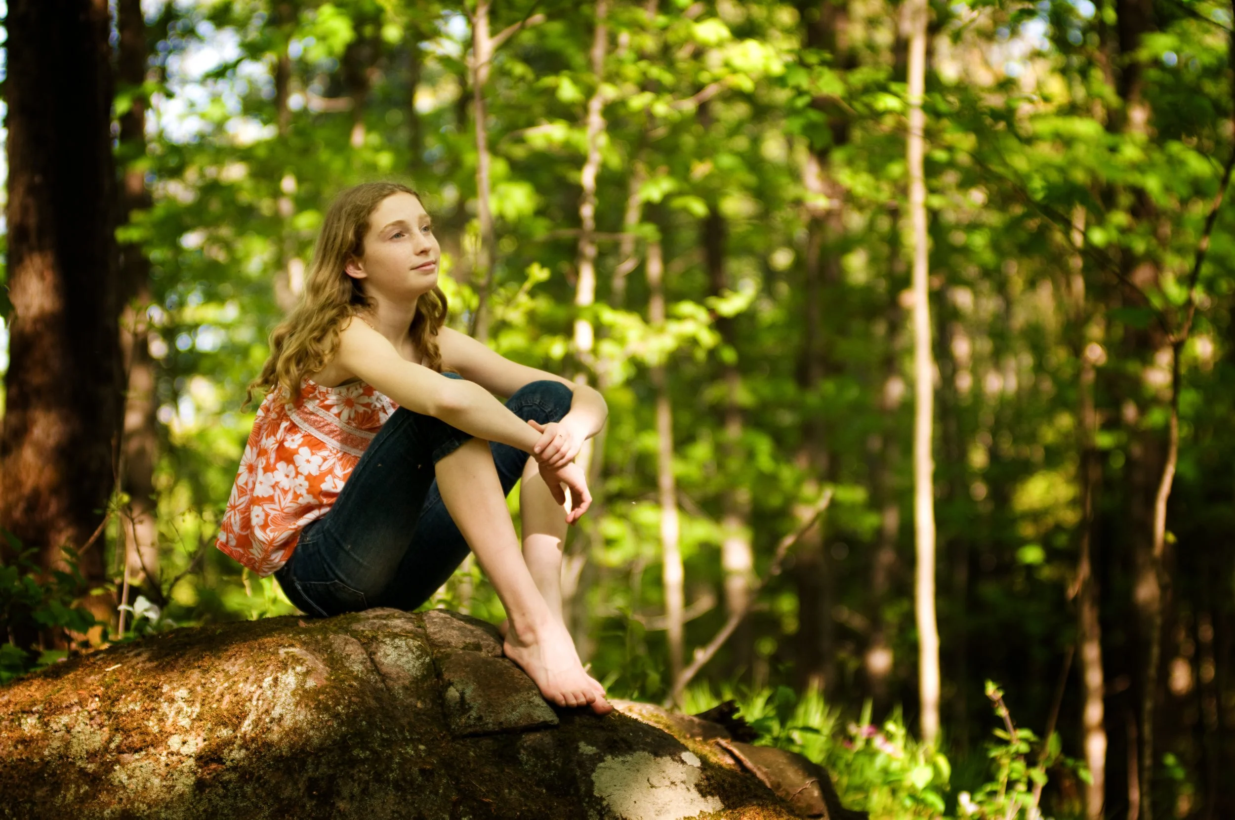 A young woman with long curly hair sits on a large moss-covered rock in a forest, wearing a sleeveless orange and white floral top and dark jeans, gazing thoughtfully into the distance.