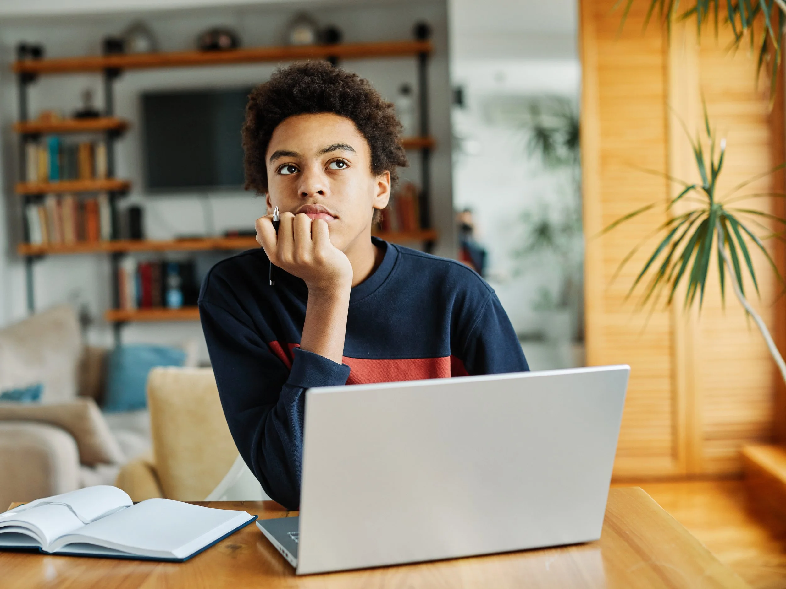 A young person sitting at a wooden table in a cozy room, with an open notebook, a laptop, and a pen, resting their chin on their hand and looking thoughtfully away.