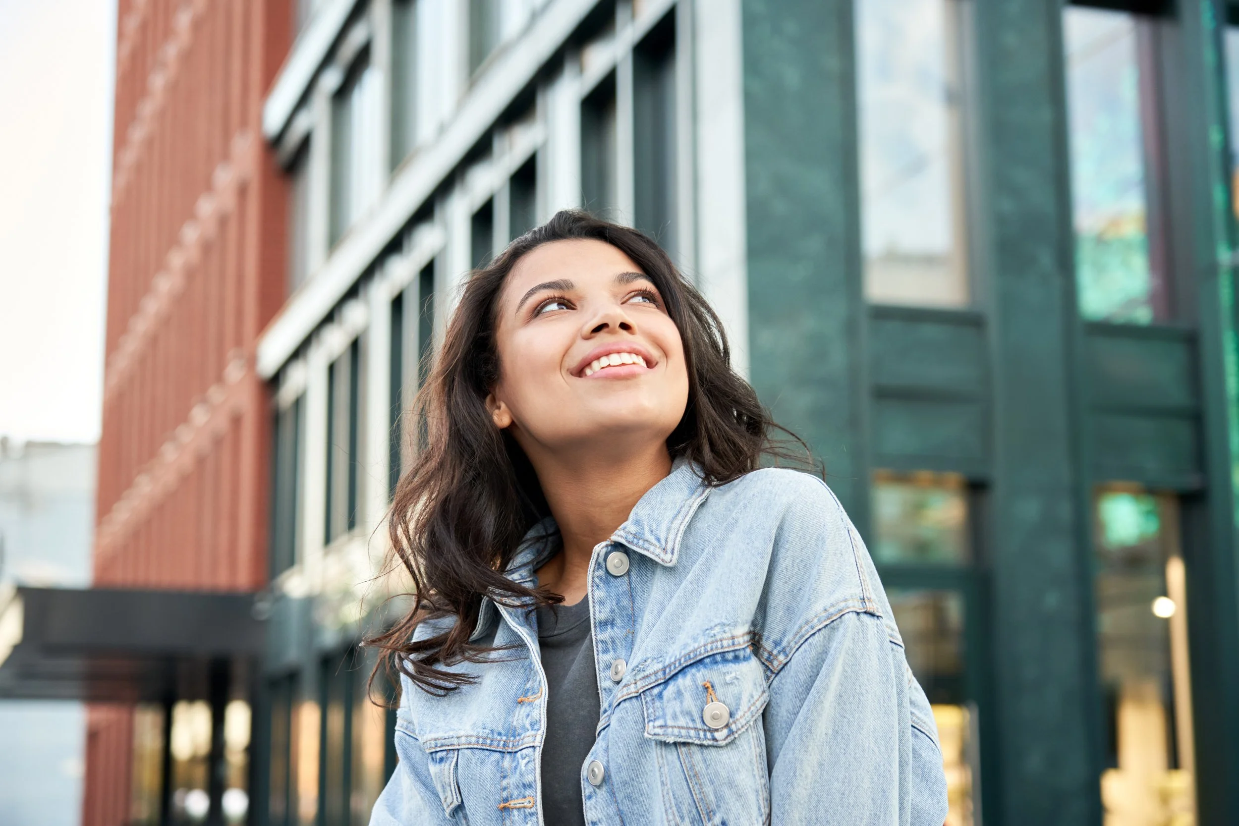 A young woman with wavy dark hair, smiling and looking upwards, wearing a denim jacket, standing outside in front of modern buildings with large windows.