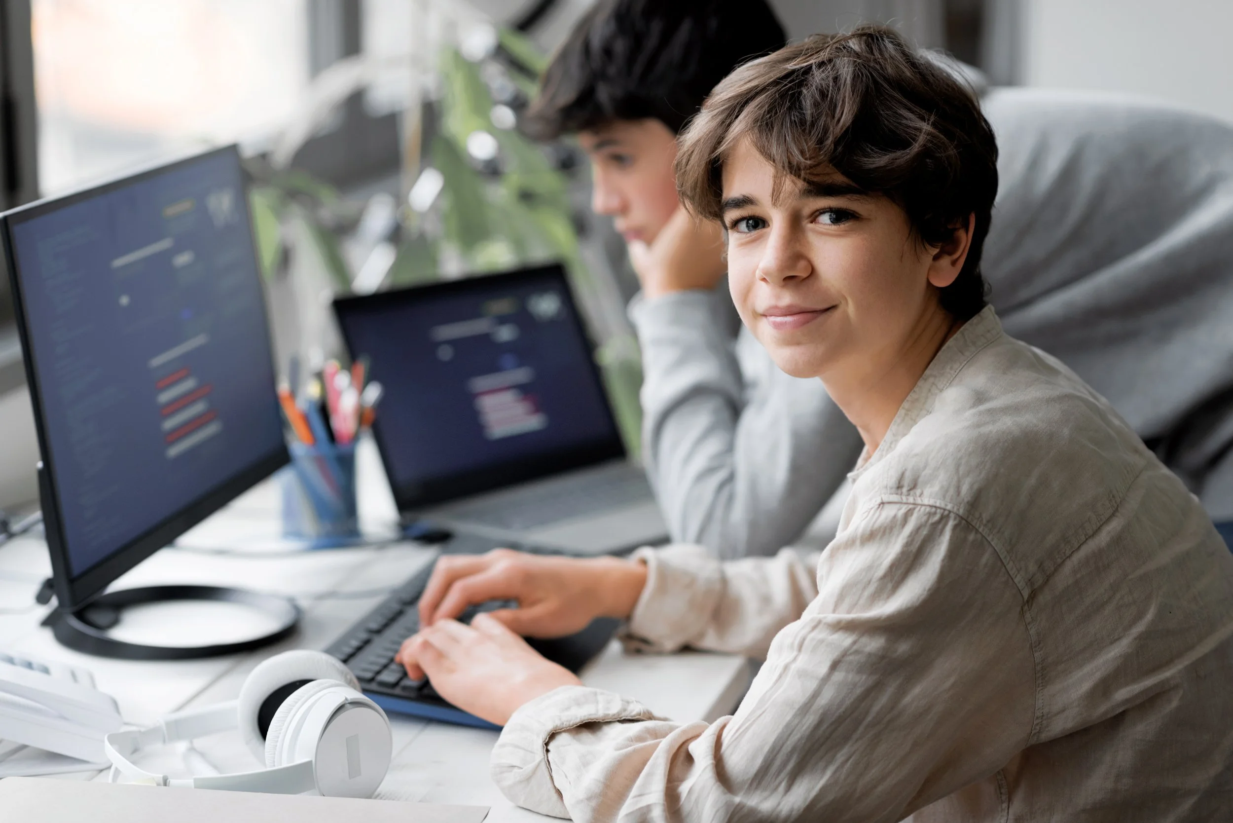 Two young boys working at a desk with computers, one smiling at the camera, the other focused on the screen, with headphones on the desk.