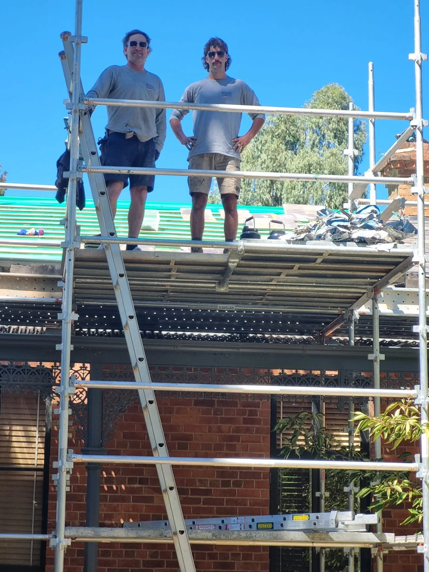 Two men standing on scaffolding on a construction site, wearing gray shirts and shorts, with a clear blue sky and trees in the background.