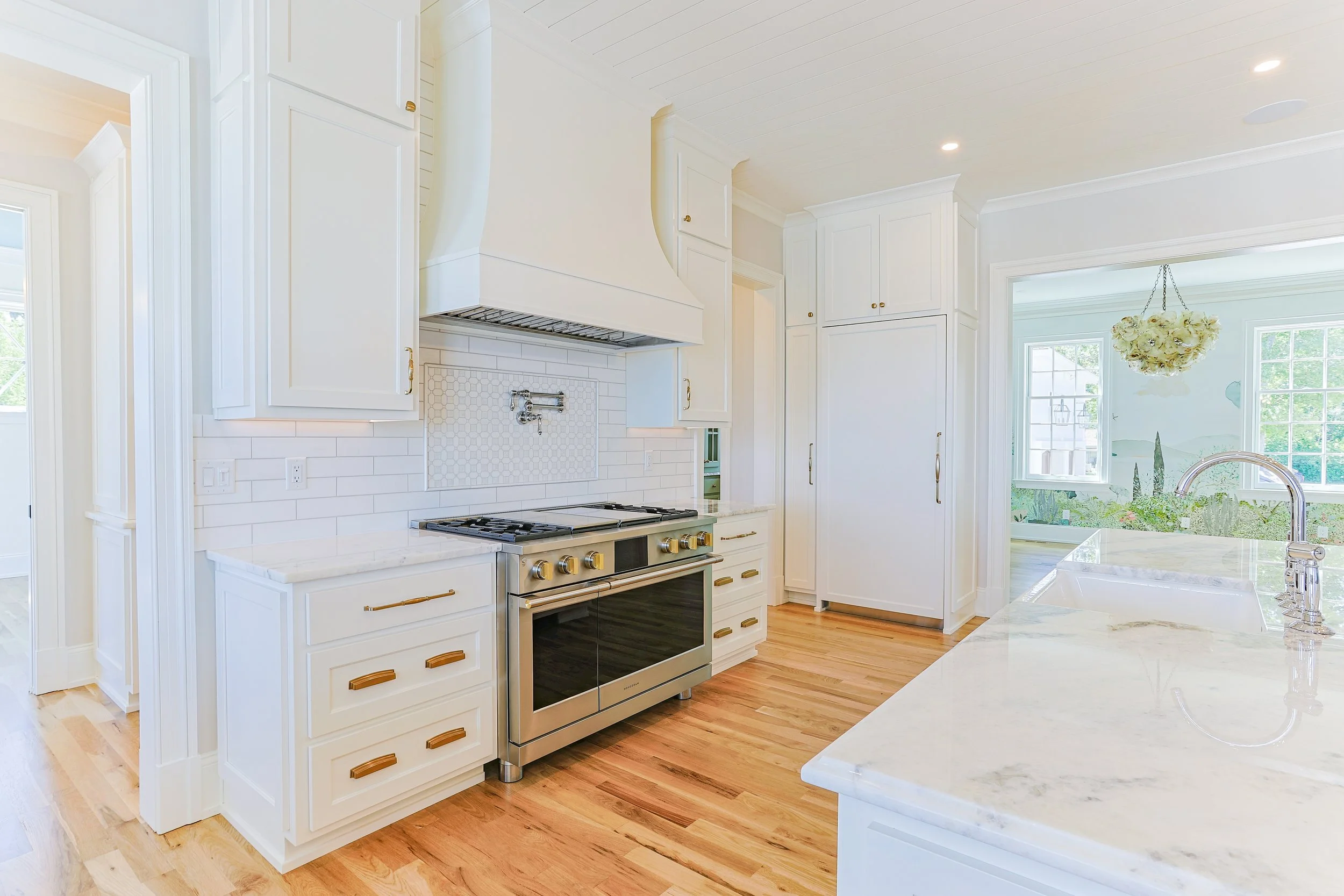 Bright kitchen with white cabinets, a gas stove, marble countertops, a double sink, and a view of a sunlit garden through large windows.
