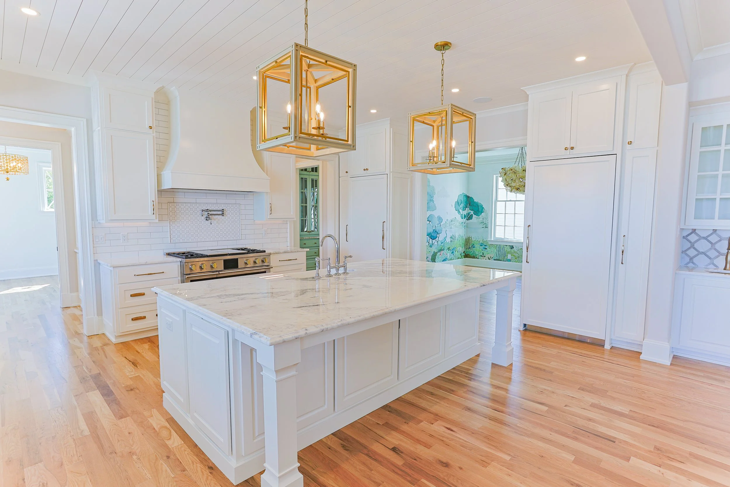 Bright white kitchen with a large island, marble countertop, gold light fixtures, wood flooring, and white cabinetry.