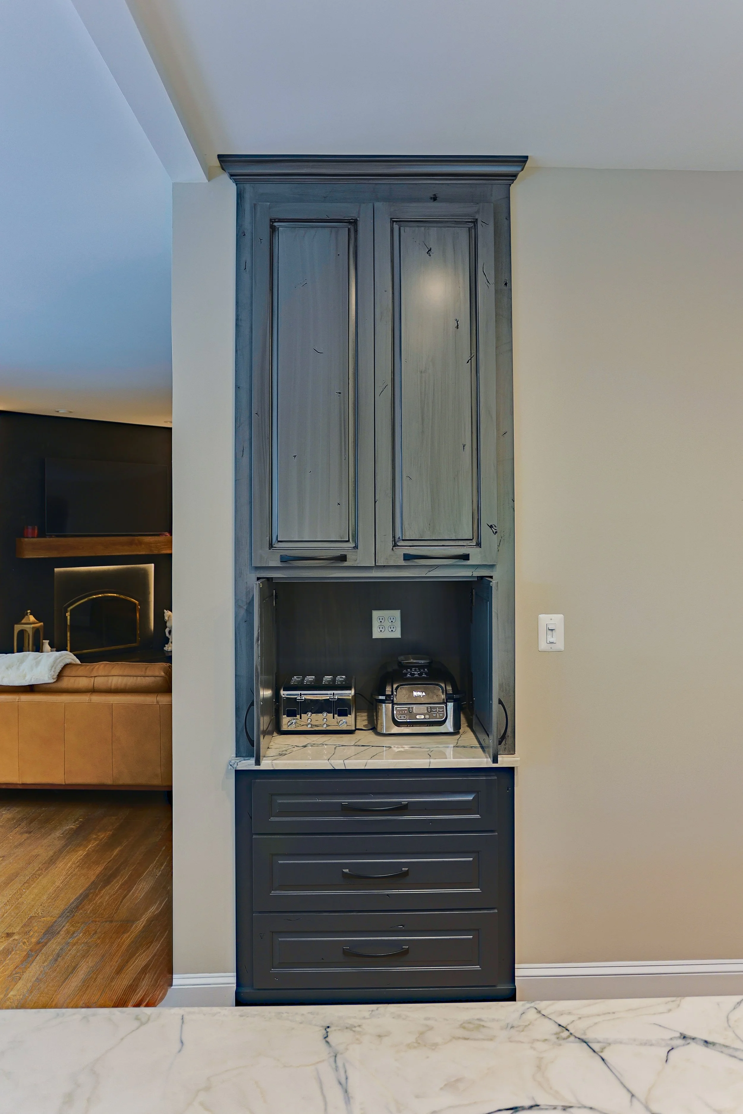 Kitchen cabinet with a marble countertop holding a toaster, a multi-cooker, and some electrical outlets, with a beige wall and a glimpse of a living room in the background.