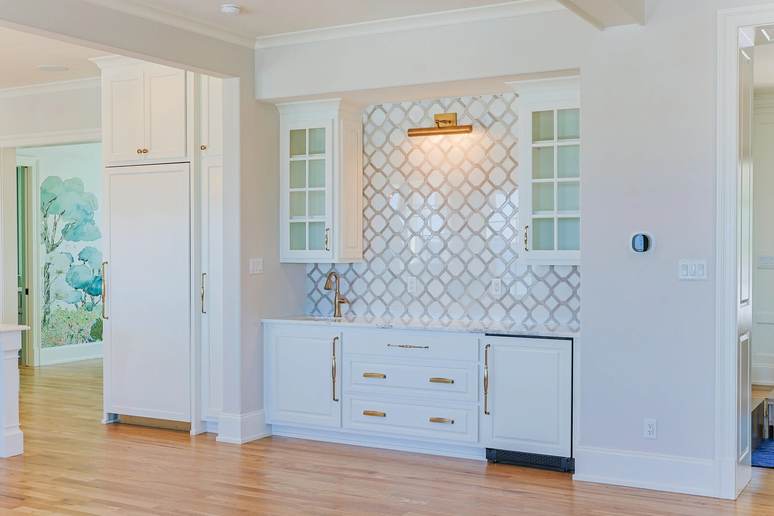 Empty kitchen with white cabinets, gold handles, patterned backsplash, and wooden floor.