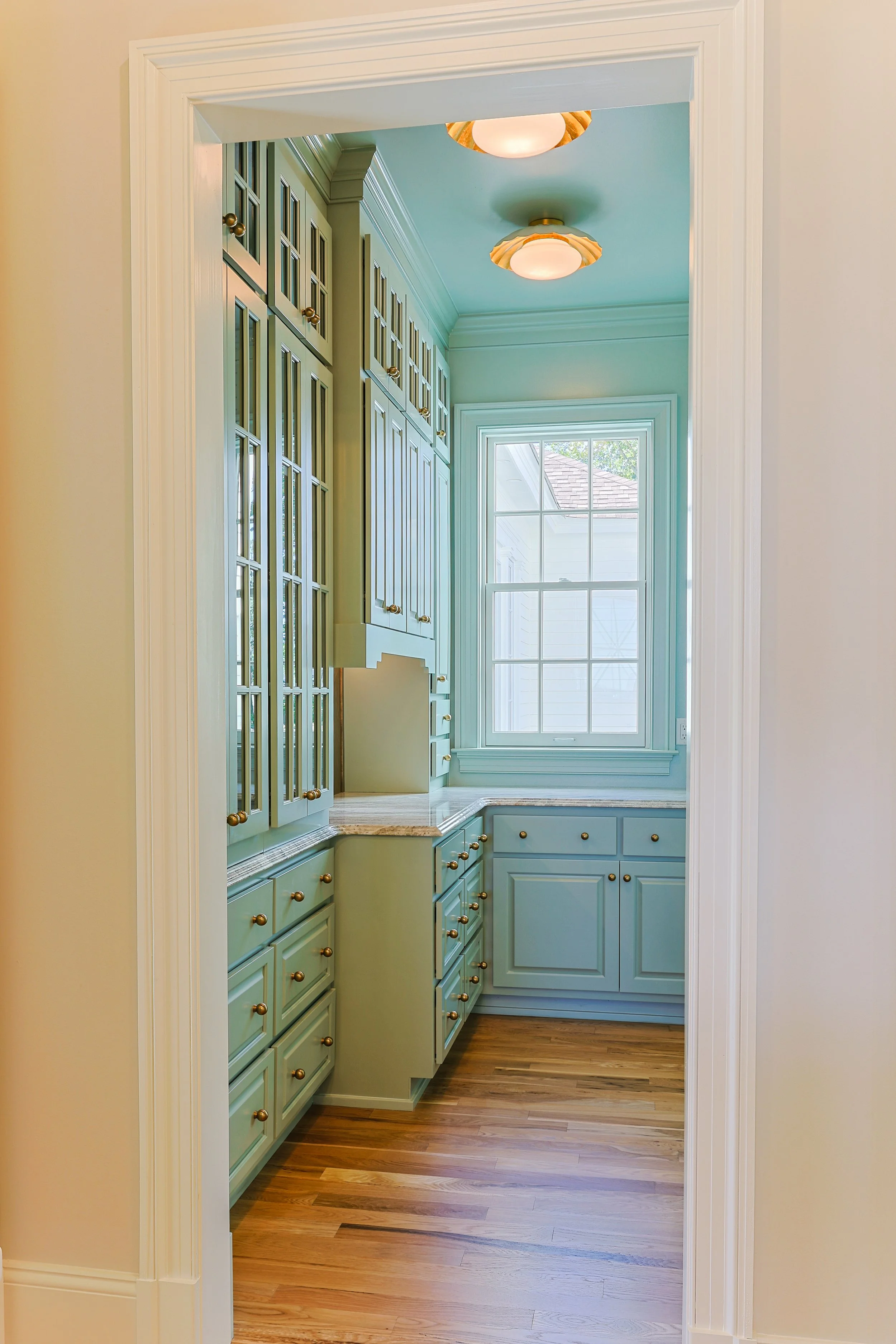 A small kitchen with mint green cabinets, hardwood floors, and a large window with a white frame. There are two ceiling lights with decorative shades.