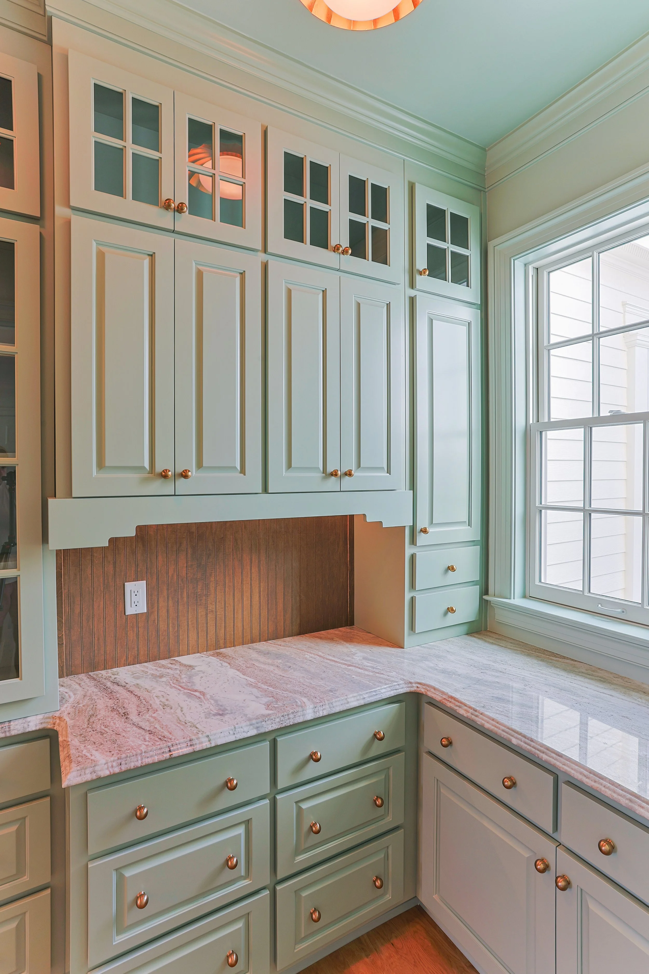 Cream-colored kitchen cabinets with gold knobs and glass-paneled upper doors, a countertop with pink and white marble pattern, and a large window letting in natural light.