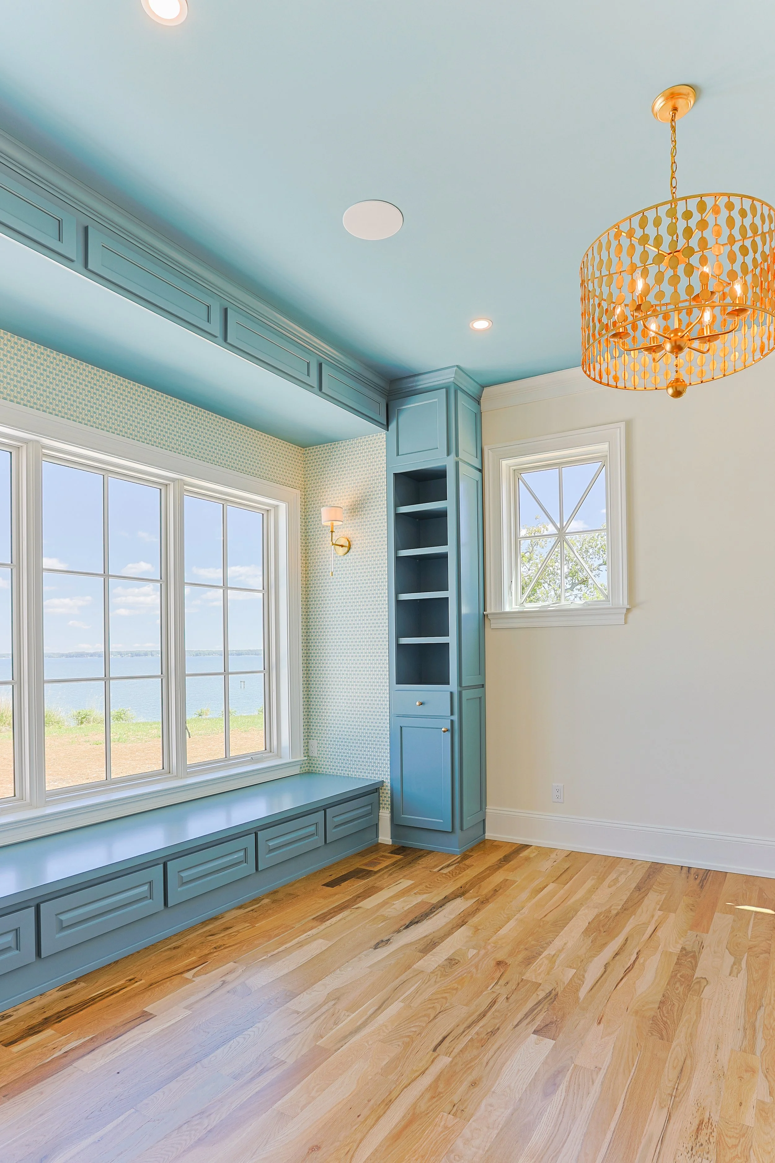 A room with light blue painted ceiling and built-in cabinets, a large window overlooking water, hardwood flooring, and a gold chandelier.