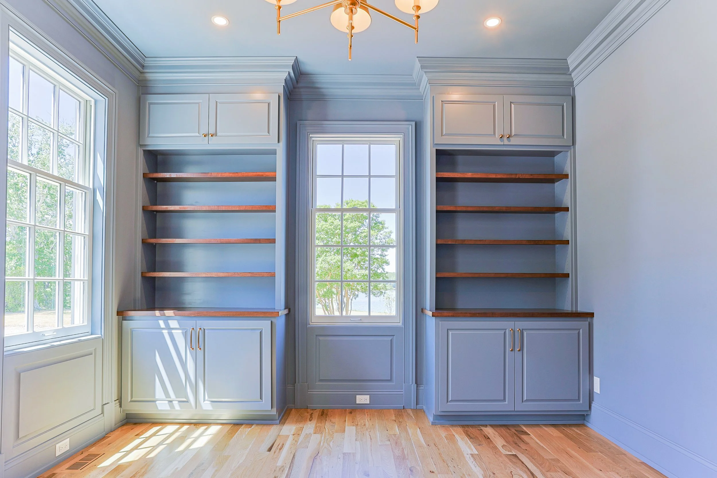 Built-in blue cabinets with open wooden shelves and gold hardware, a central window with a view of trees outdoors, a hardwood floor, and a ceiling light fixture.