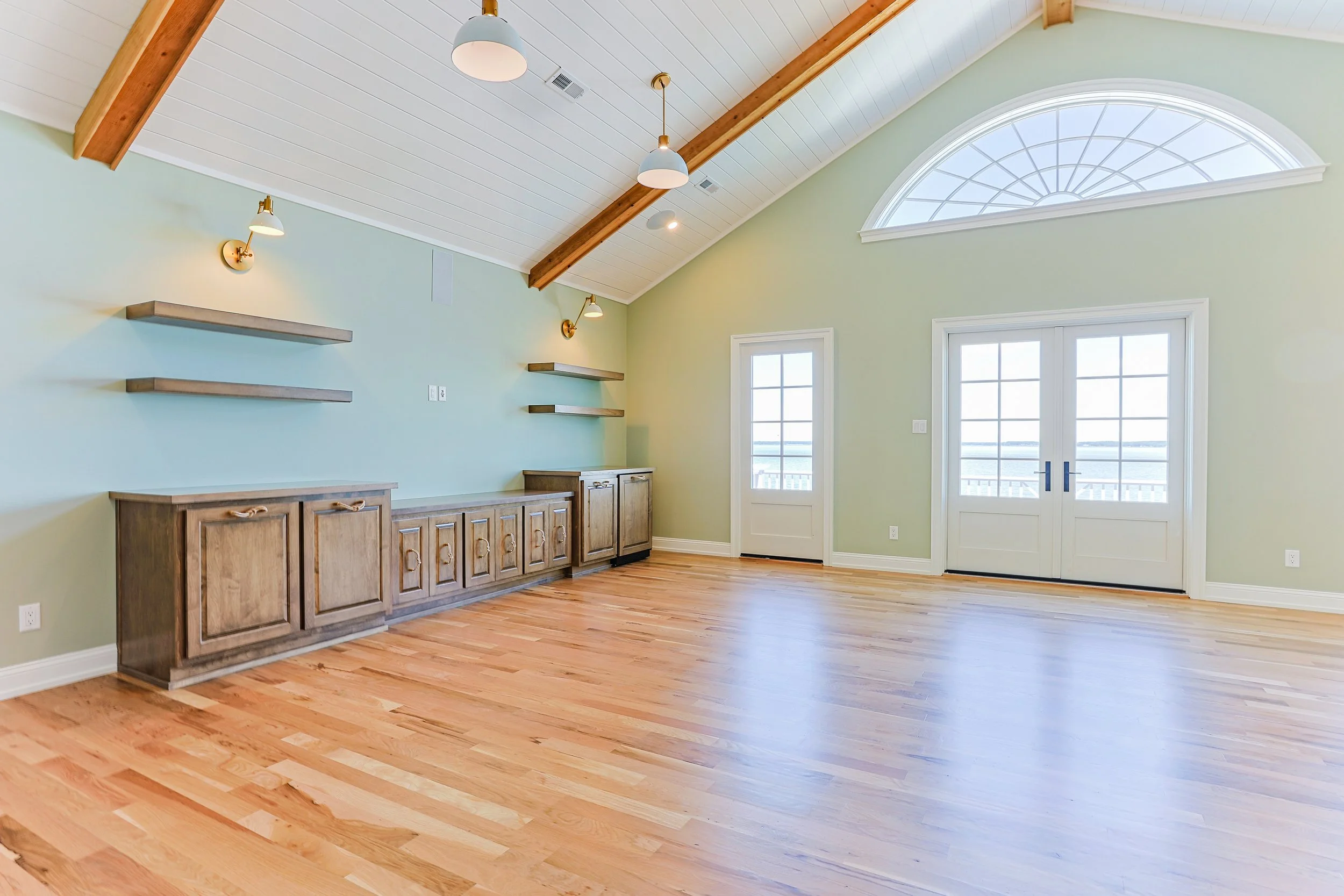 Spacious room with light green walls, wooden ceiling, hardwood floor, large windows, and double glass doors leading outside.