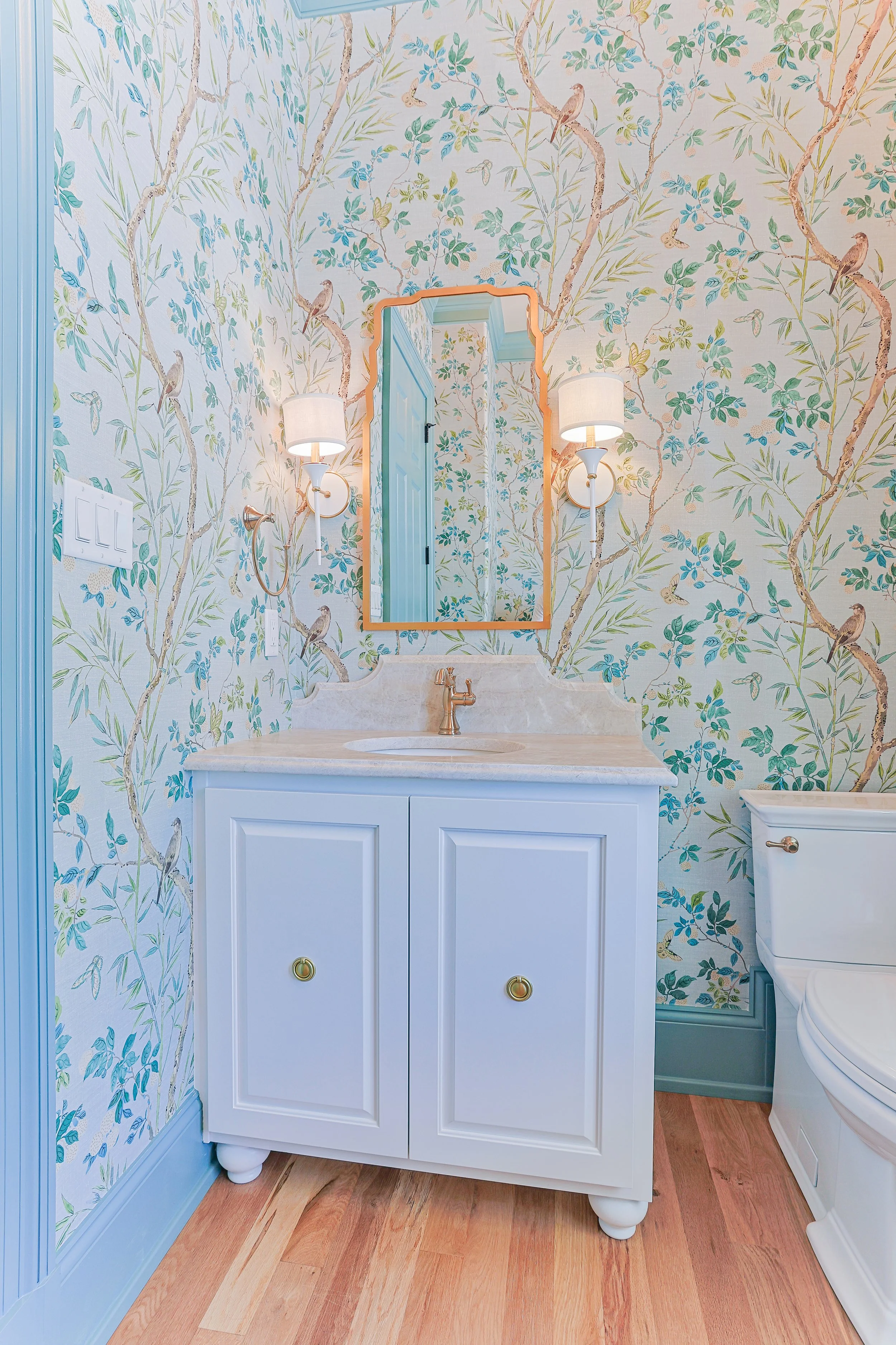 A bathroom with wallpaper featuring birds and branches, a white vanity with a marble top, a mirror with a decorative frame, and wall sconces with white shades.