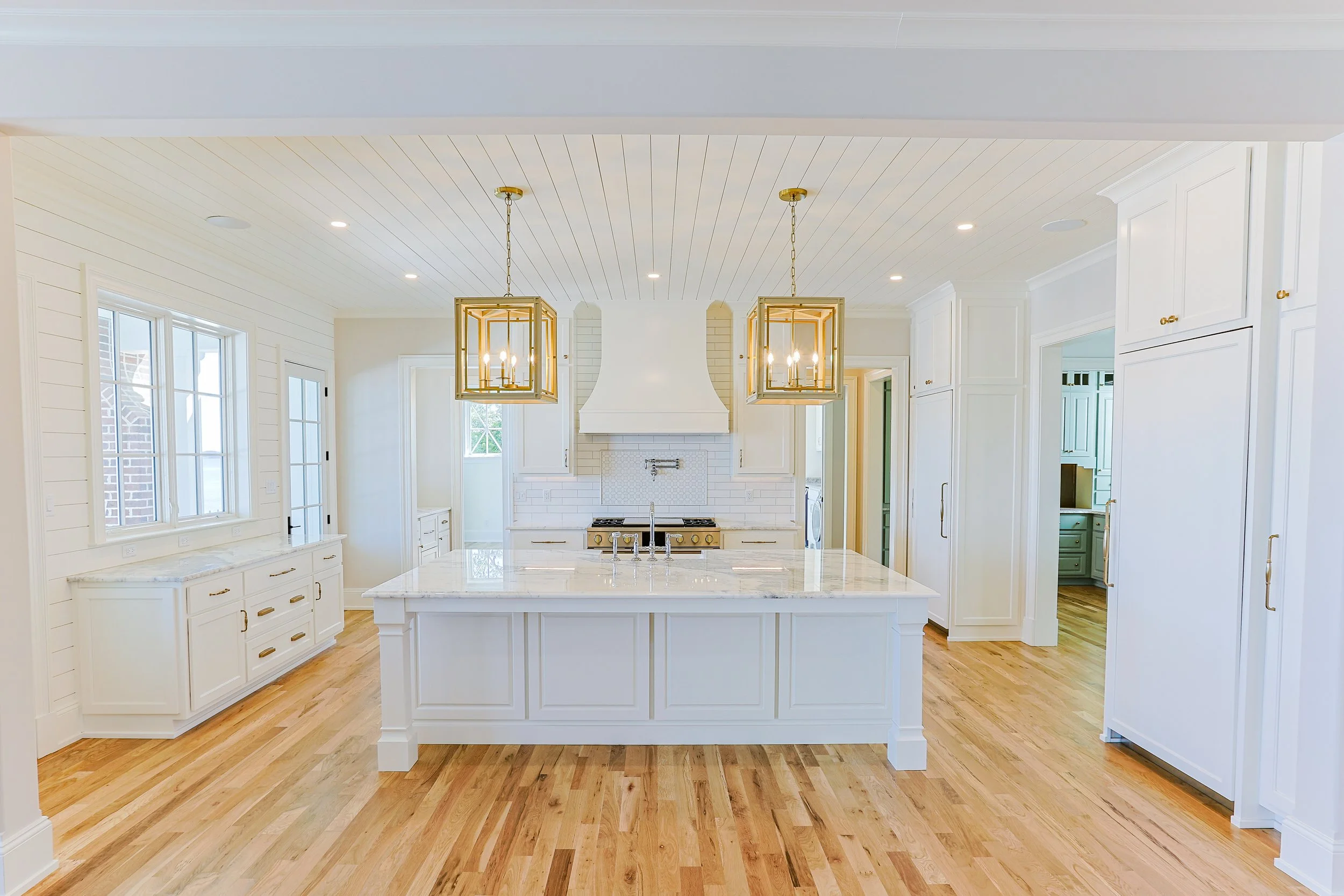 Bright, modern kitchen with white cabinetry, a central island with marble countertop, gold hardware, and two gold pendant lights. Hardwood floors and a white brick range hood with a stove underneath.
