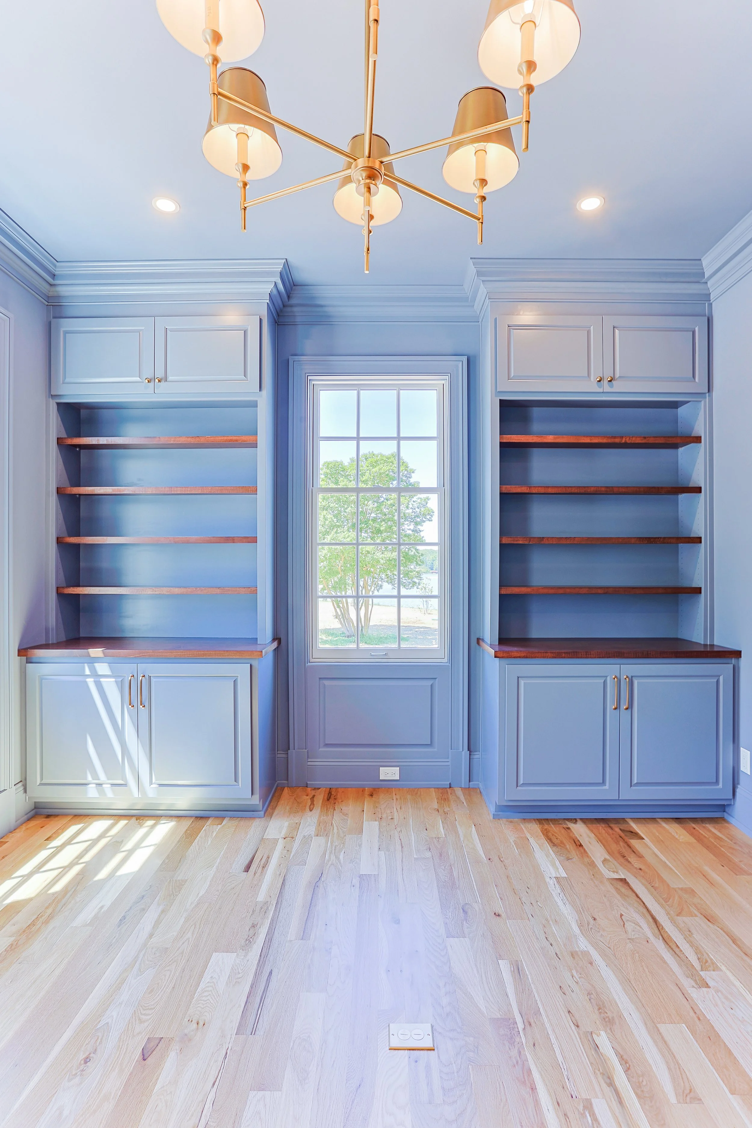 A bright blue built-in bookshelf with wooden shelves and gold handles, centered around a window with a view of a tree outside, inside a room with hardwood flooring and a modern chandelier.