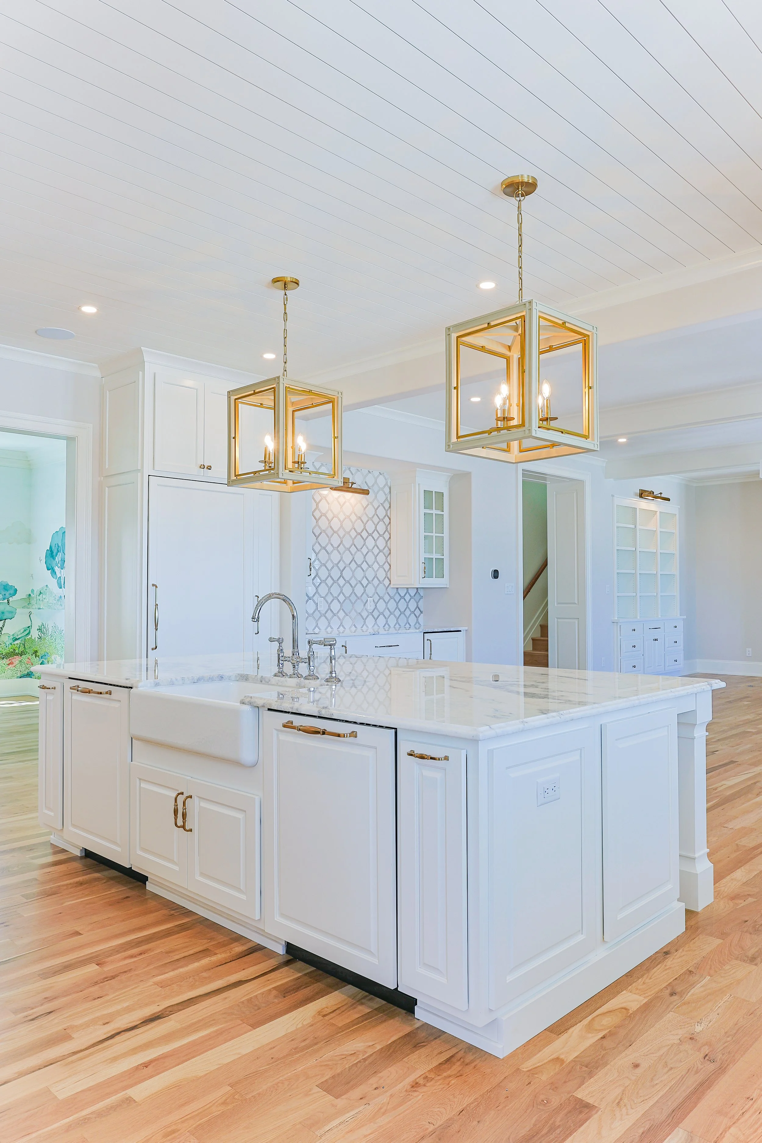 Bright, modern kitchen with white cabinets, a white marble island, and gold light fixtures hanging from a white ceiling, with light wood flooring.