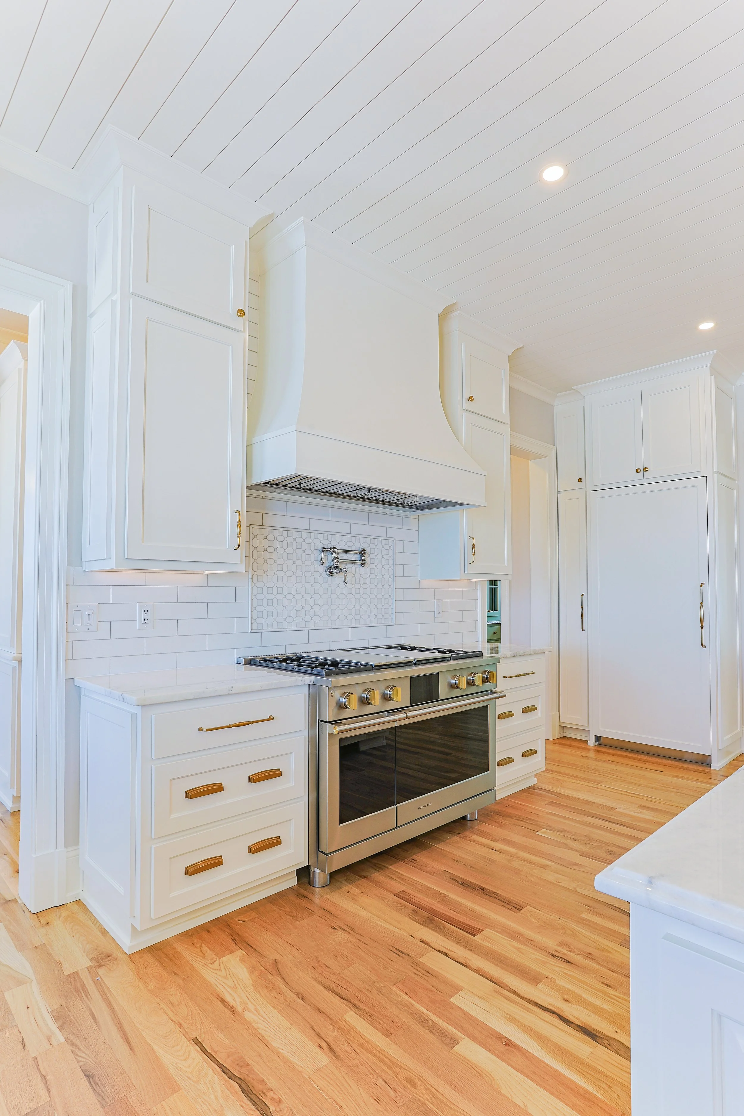 Bright white kitchen with wooden floors, cabinets, and a stainless steel oven, stove, and vent hood.