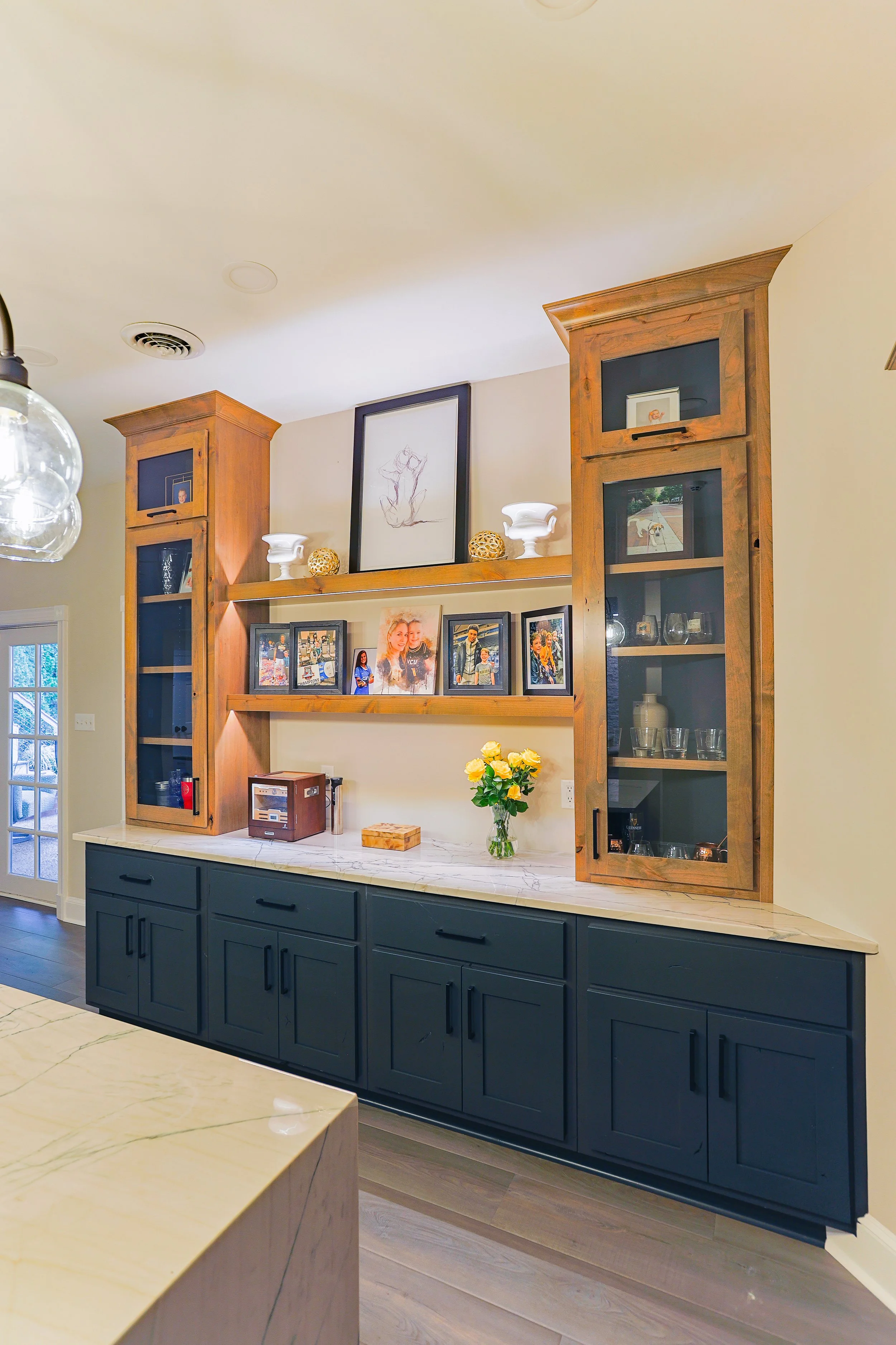 A built-in wooden cabinet with glass doors, filled with glassware, above a marble countertop with a vase of yellow roses, framed photos, and decorative items, in a bright room with a window.
