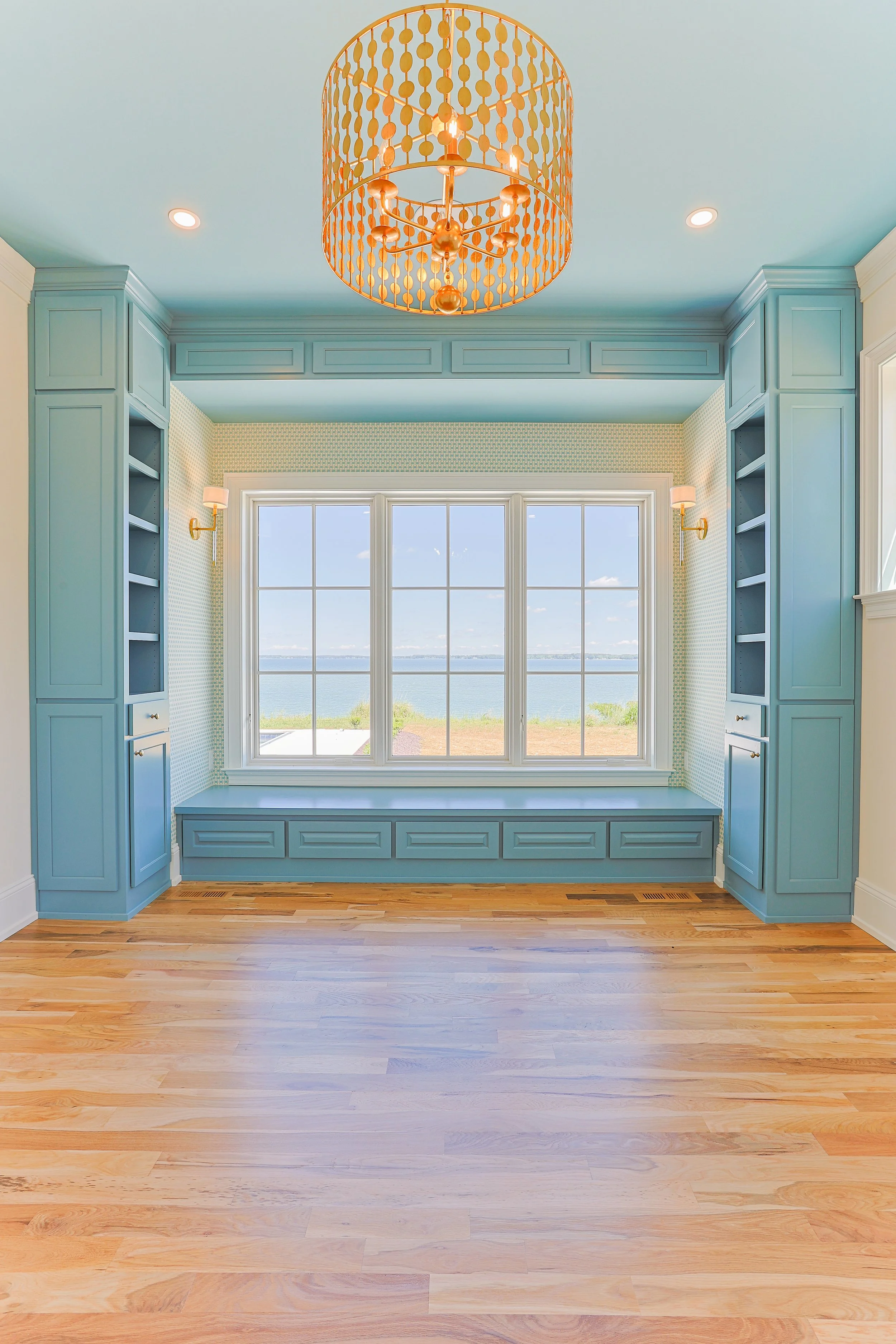 A bright room with light blue walls and built-in blue cabinetry on either side of a large window with a view of water and sky, hardwood flooring, and a decorative orange chandelier hanging from the ceiling.