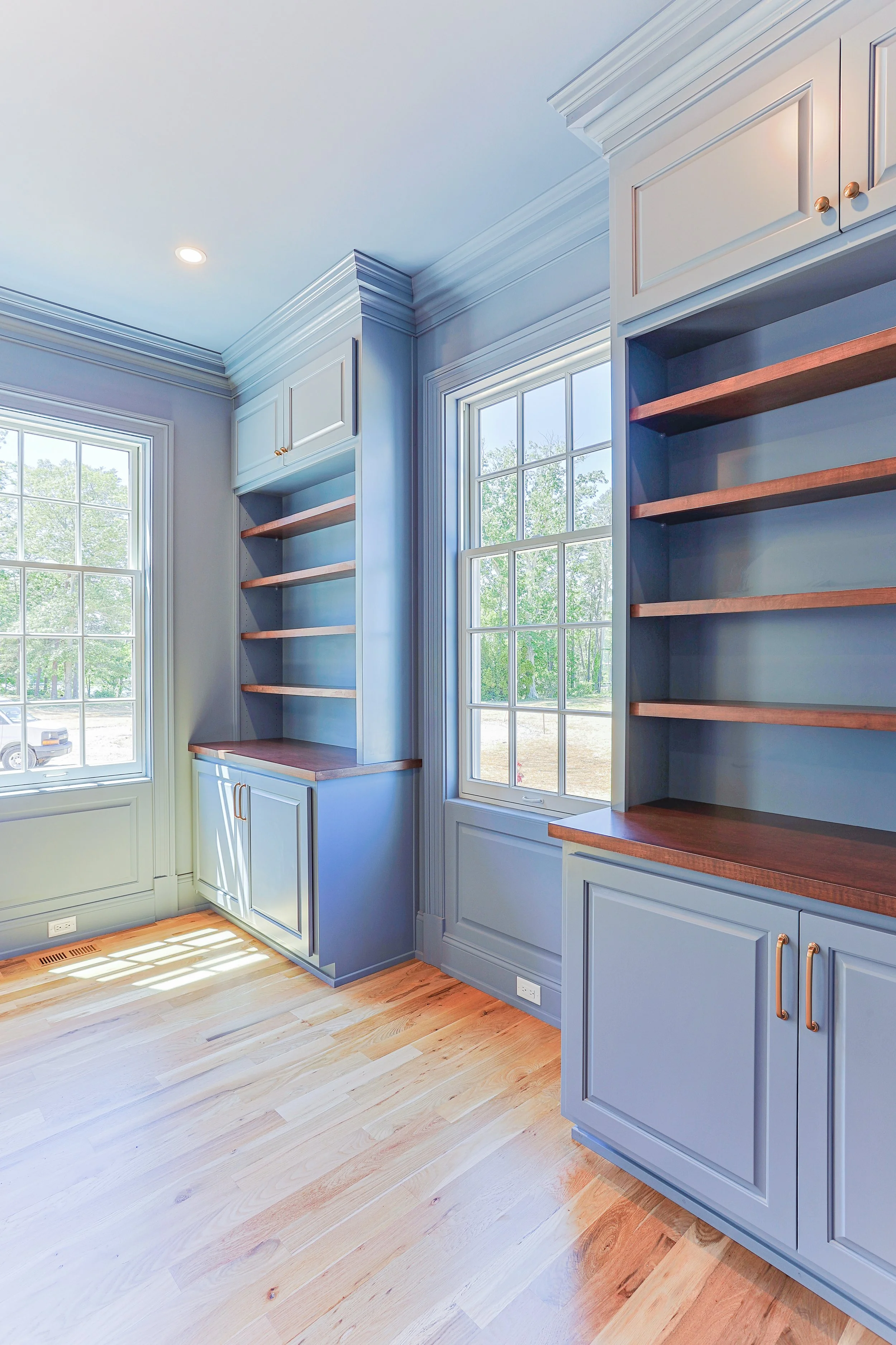 Room with large windows, built-in blue bookshelves with wooden shelves and gold handles, light-colored hardwood floor, and crown molding.