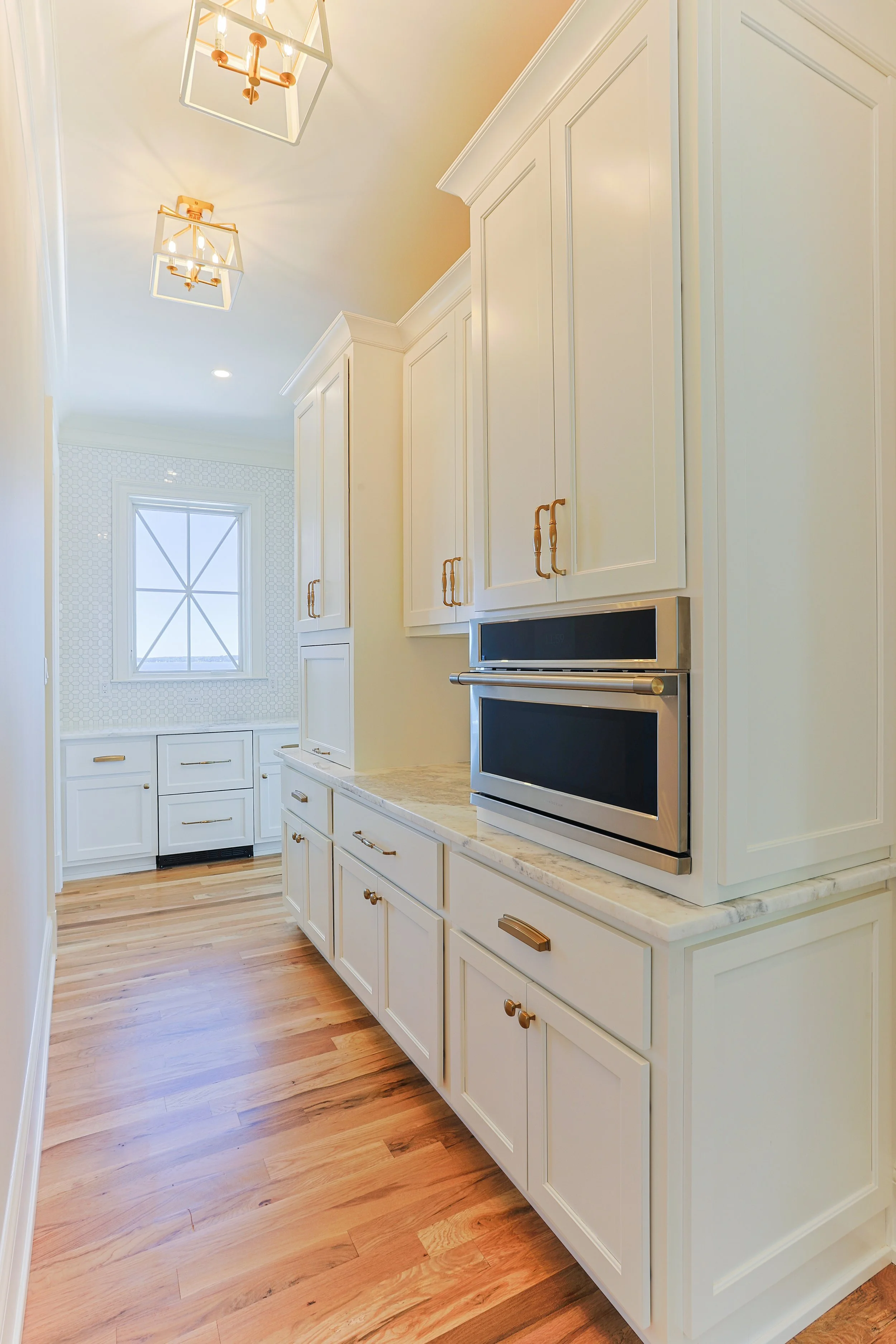 A bright, modern kitchen with white cabinets, gold hardware, and wood flooring. Stainless steel double oven built into the cabinetry and a window at the end of the room letting in natural light.
