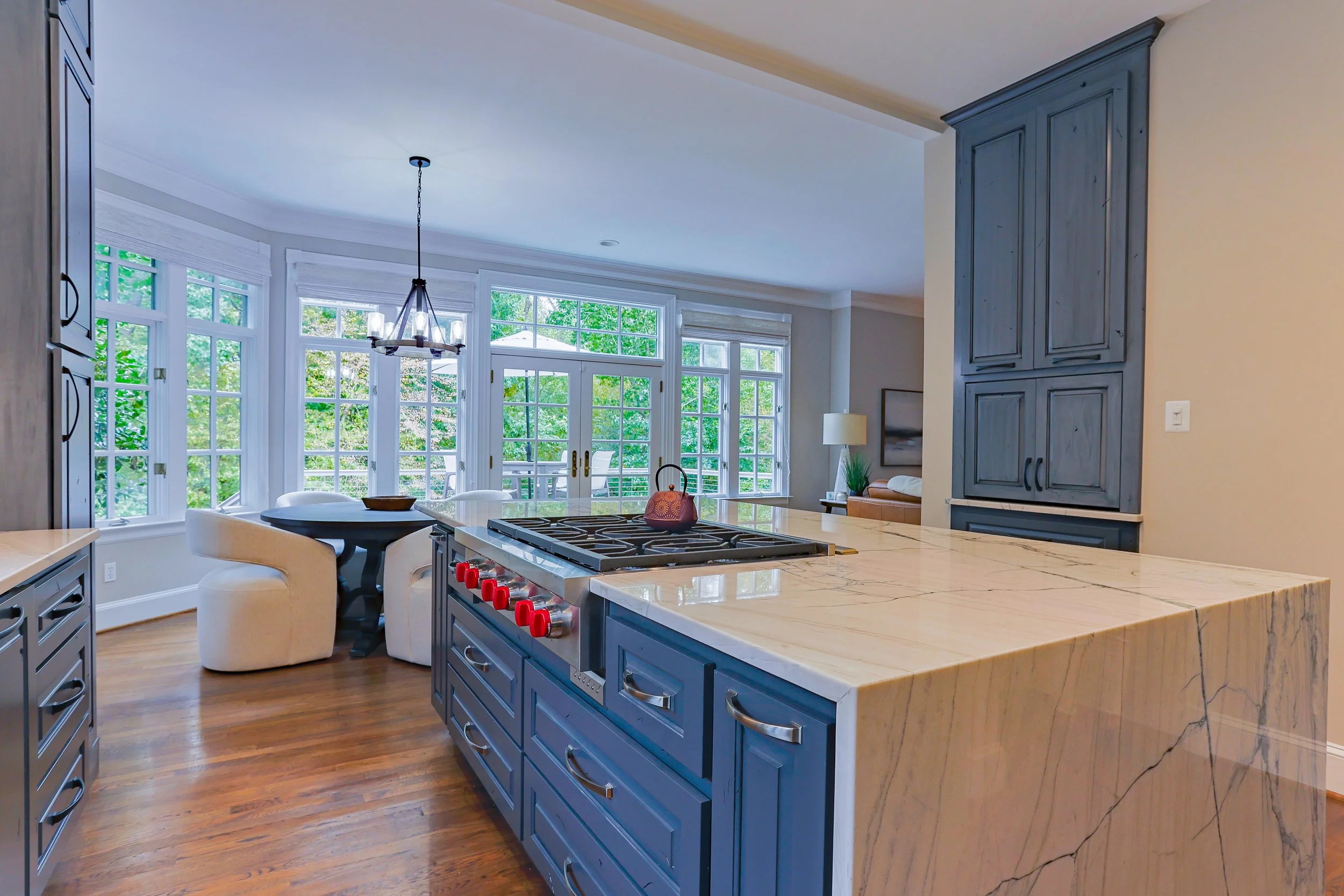 Modern kitchen with blue cabinets, marble island, and large bay windows overlooking greenery.