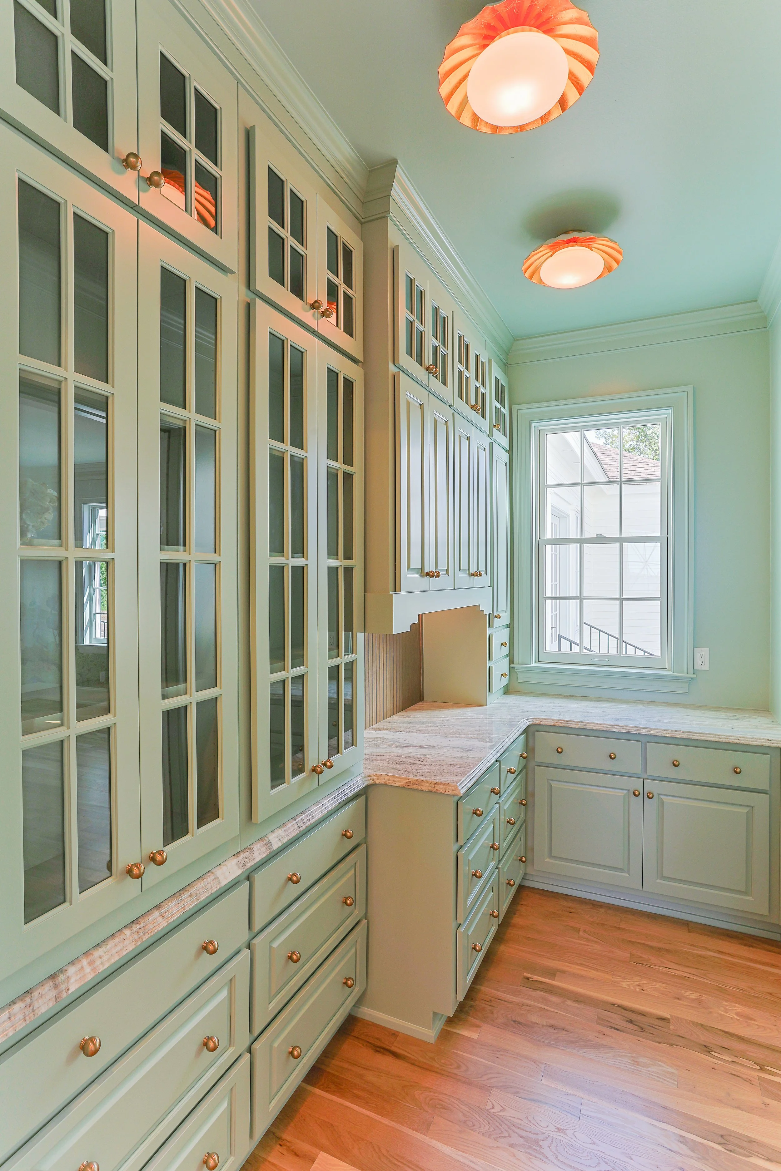 Bright kitchen with light green cabinets and wooden floors, illuminated by ceiling lights, with a large window at the end.