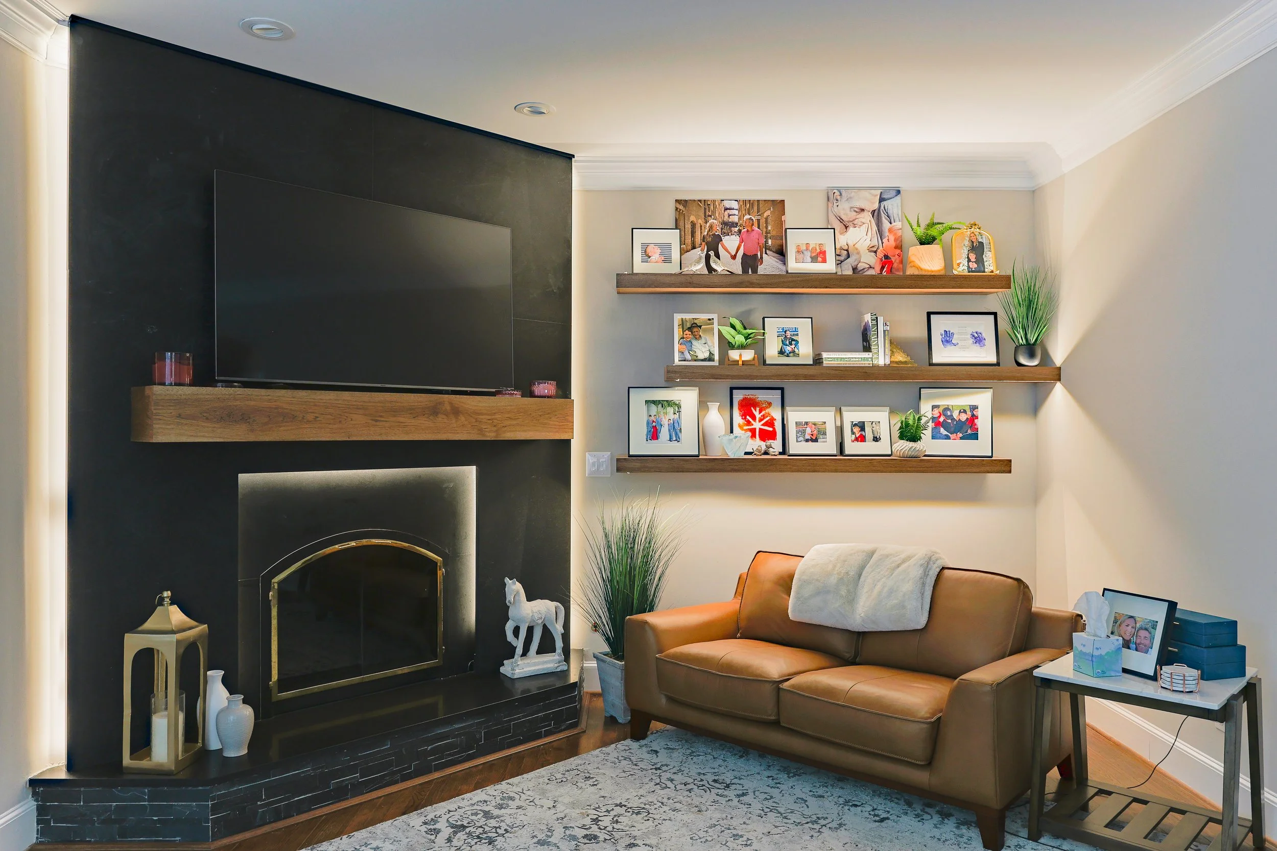 Living room featuring a black fireplace with wooden mantel, mounted flat-screen TV, tan leather sofa with a blanket, three wooden shelves with family photos, plants, and decorative items, a side table with framed photos and tissue box, and a patterne